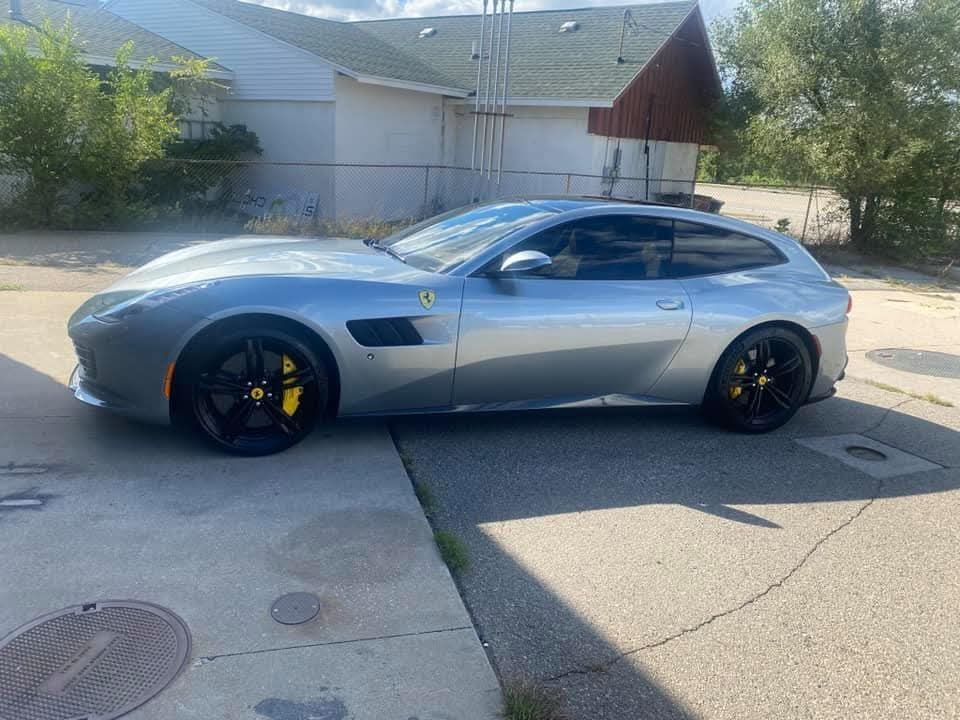 Silver Ferrari GTC4Lusso parked on a paved driveway with black wheels, yellow brake calipers, and a building in the background.