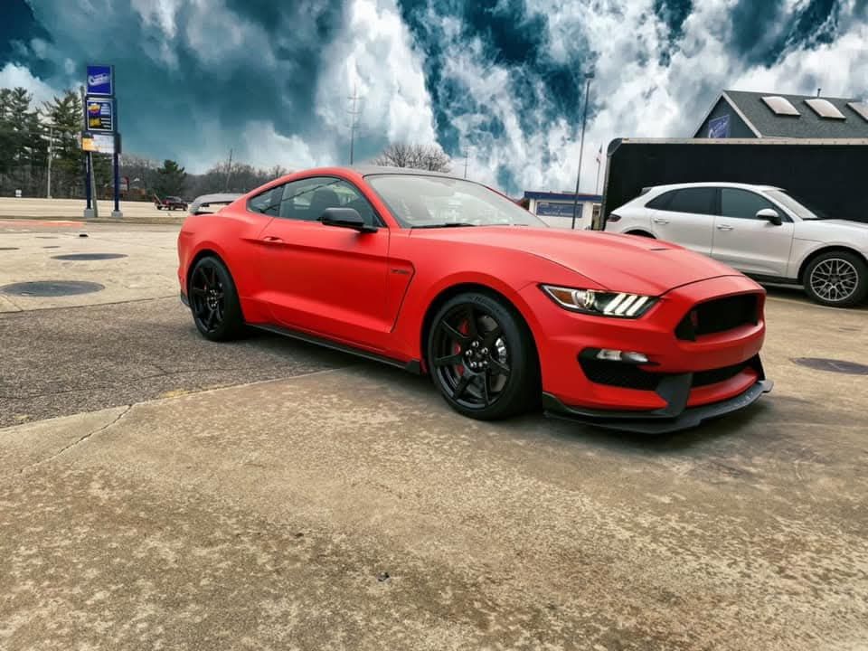 Red Ford Mustang on a concrete surface, cloudy sky in the background. Black wheels.