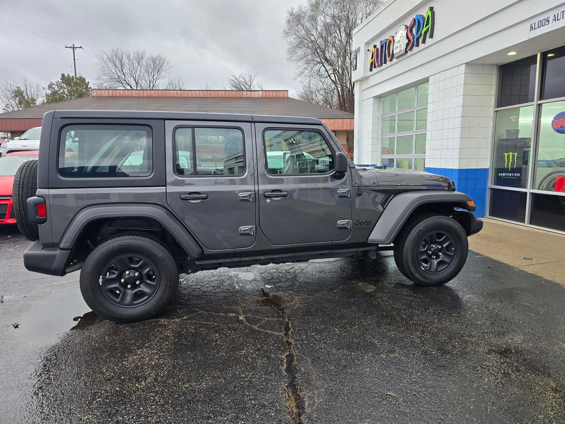 Gray Jeep Wrangler parked outside an auto shop, with black wheels, and a wet parking lot.