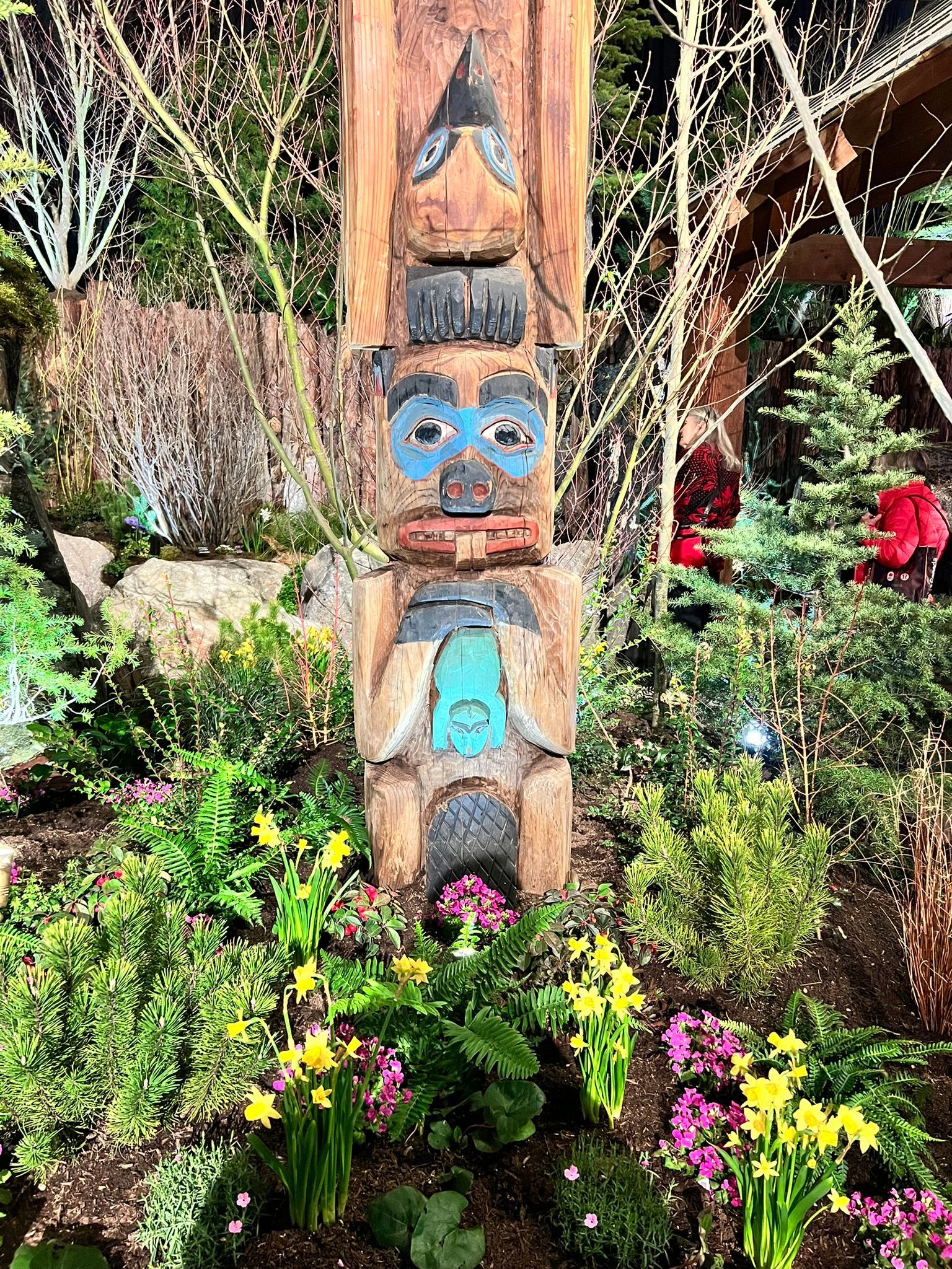 A totem pole is surrounded by flowers and trees in a garden.
