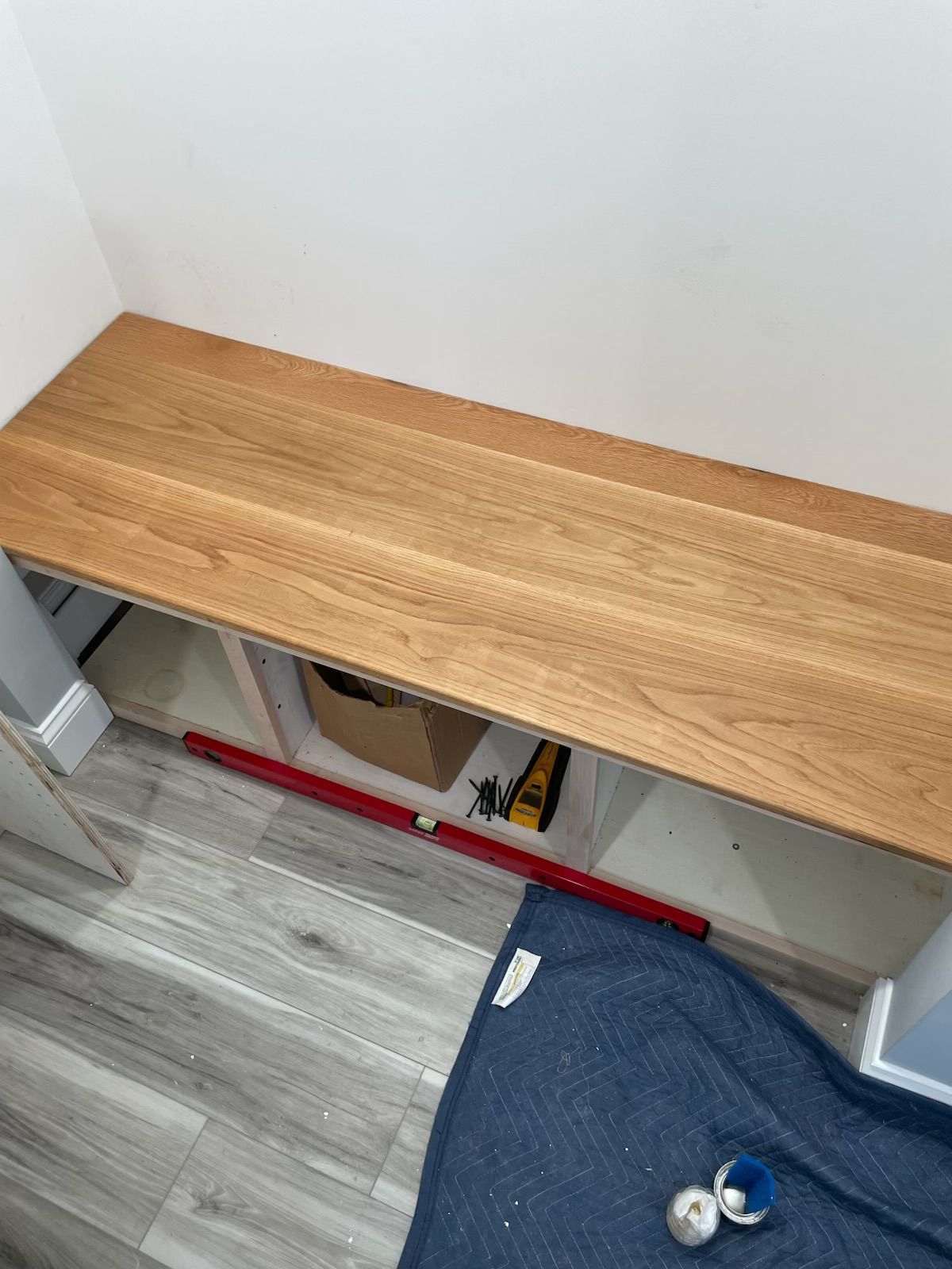 Wooden bench in a niche, holding tools and a cardboard box. A blue cushion and tools are on the floor.