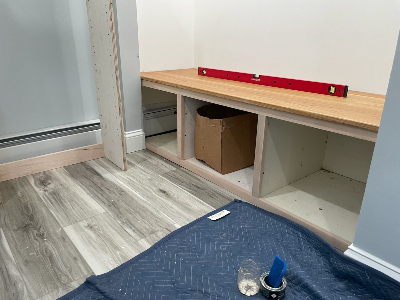 A partially built closet bench with cubby holes. A level sits on the wooden top. Cardboard box in one cubby.