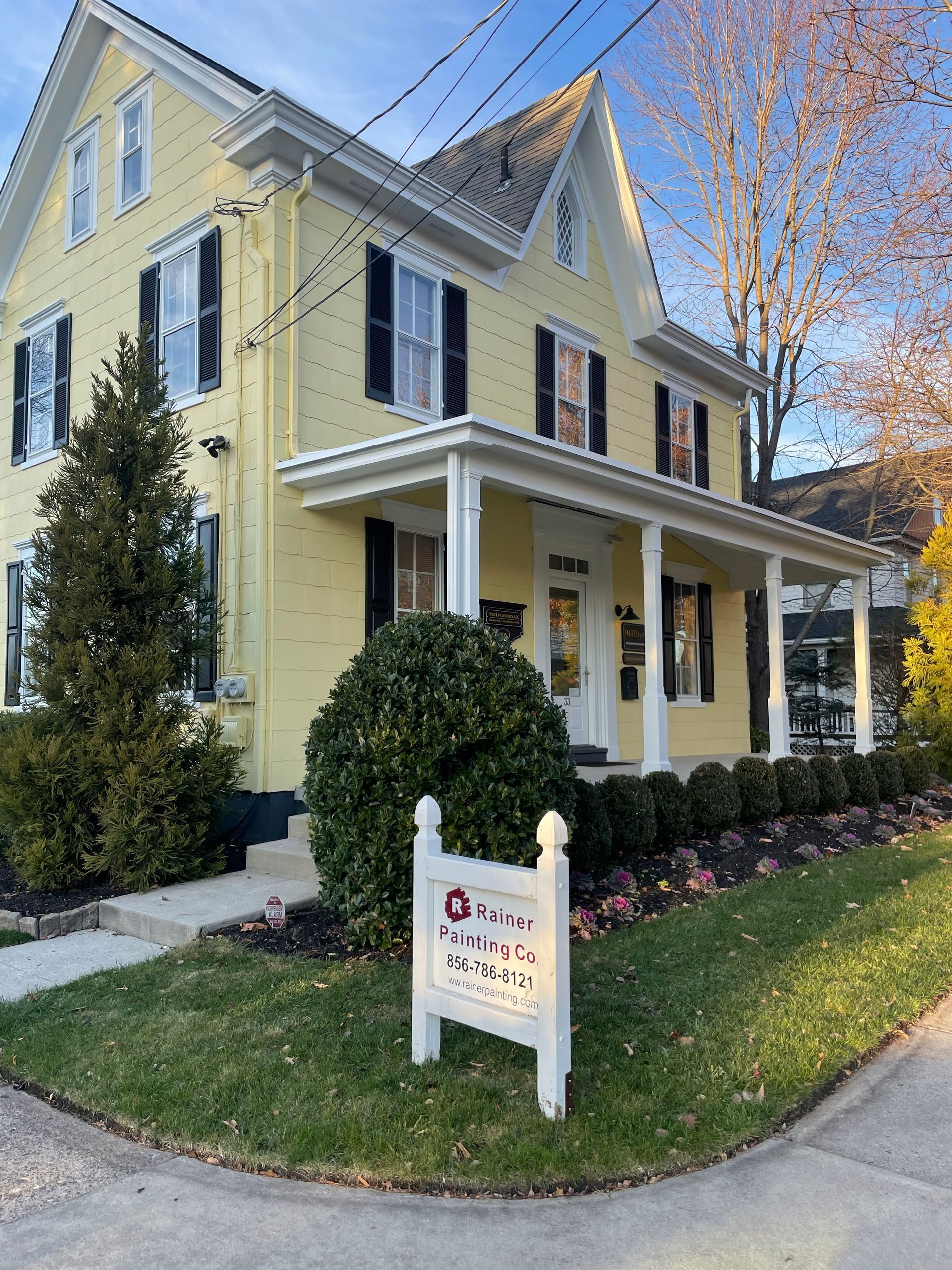 Yellow house with black shutters, a porch, and a sign in front; sunny day.