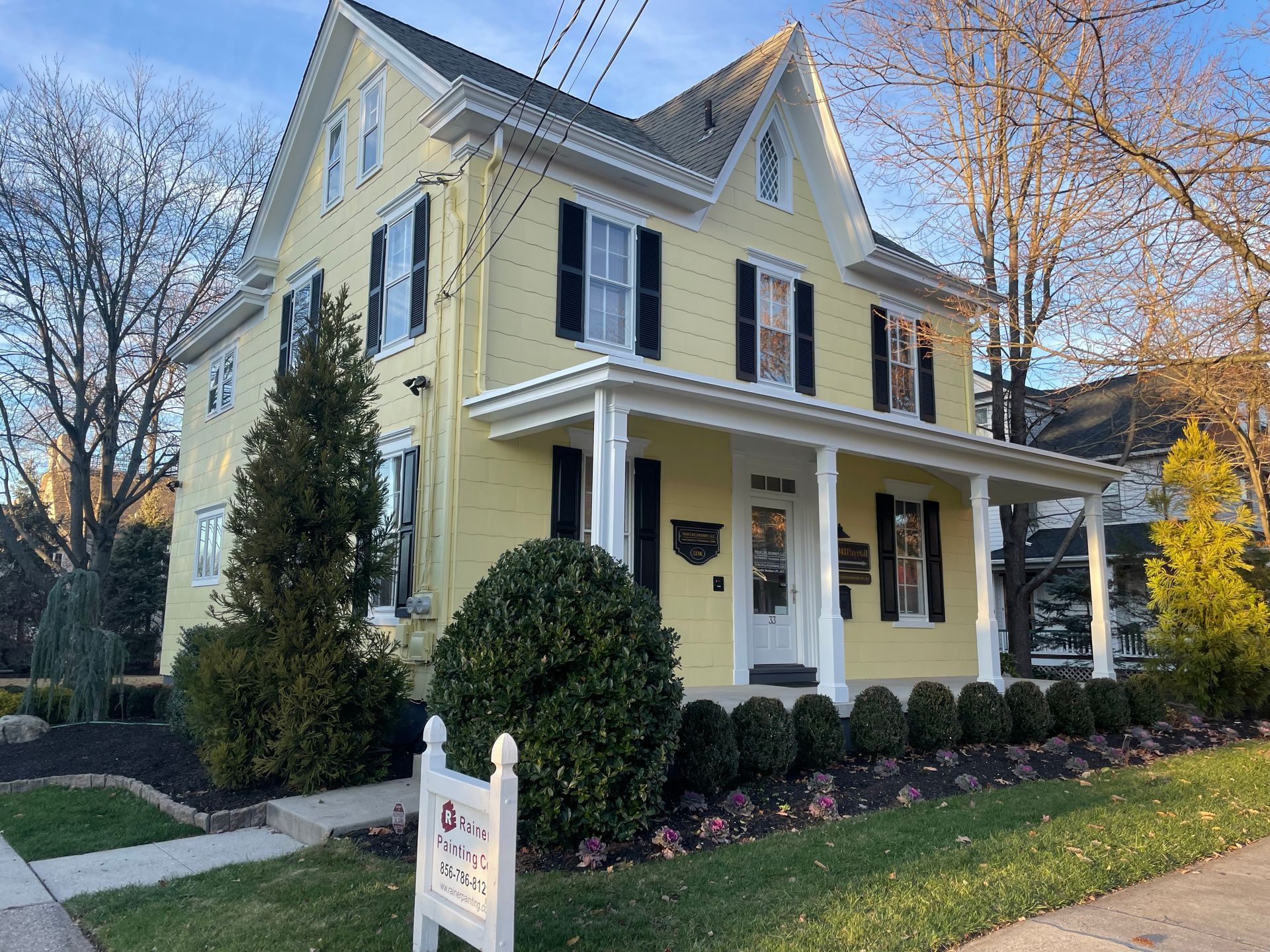 Yellow two-story house with black shutters, white trim, and a porch, set in a well-landscaped yard.
