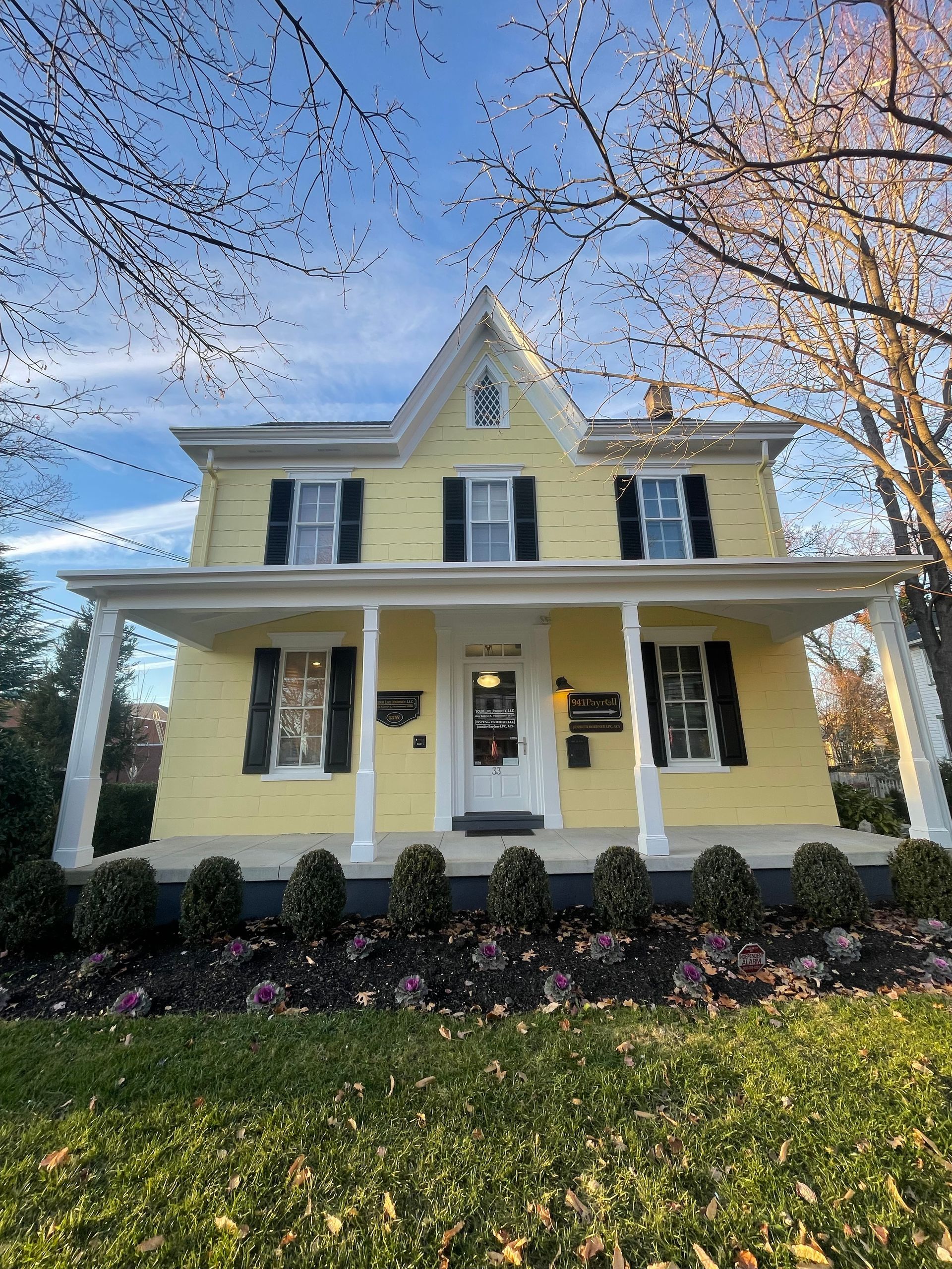 Yellow house with black shutters, white porch, and landscaping under a blue sky.