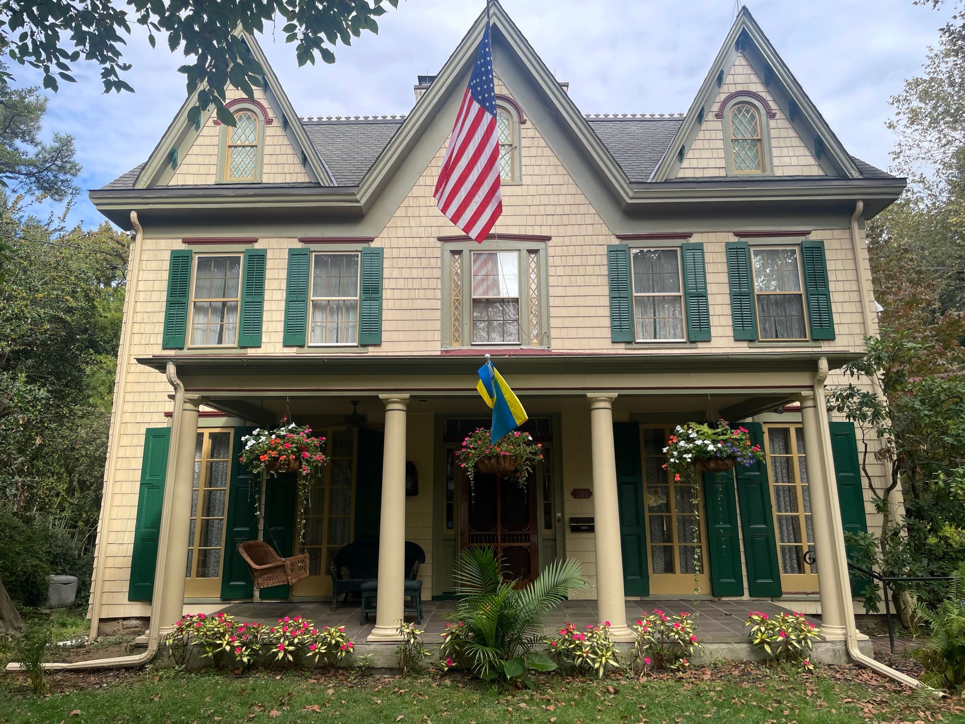 Two-story house with green shutters, a porch, American and Ukrainian flags. Flowers decorate the porch; trees surround the house.