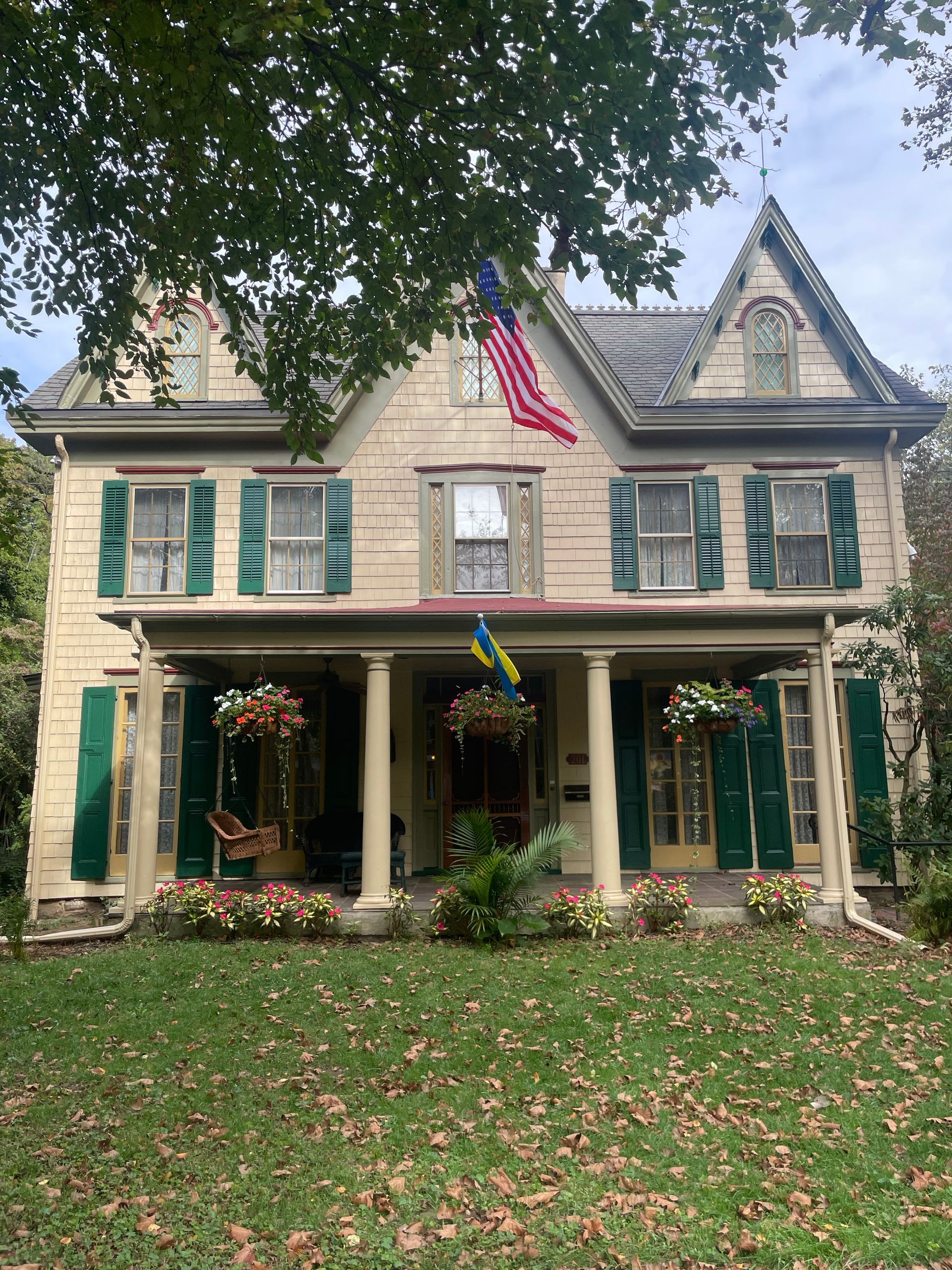 Two-story house with green shutters, a porch, American flag, flowers, and a grassy lawn.