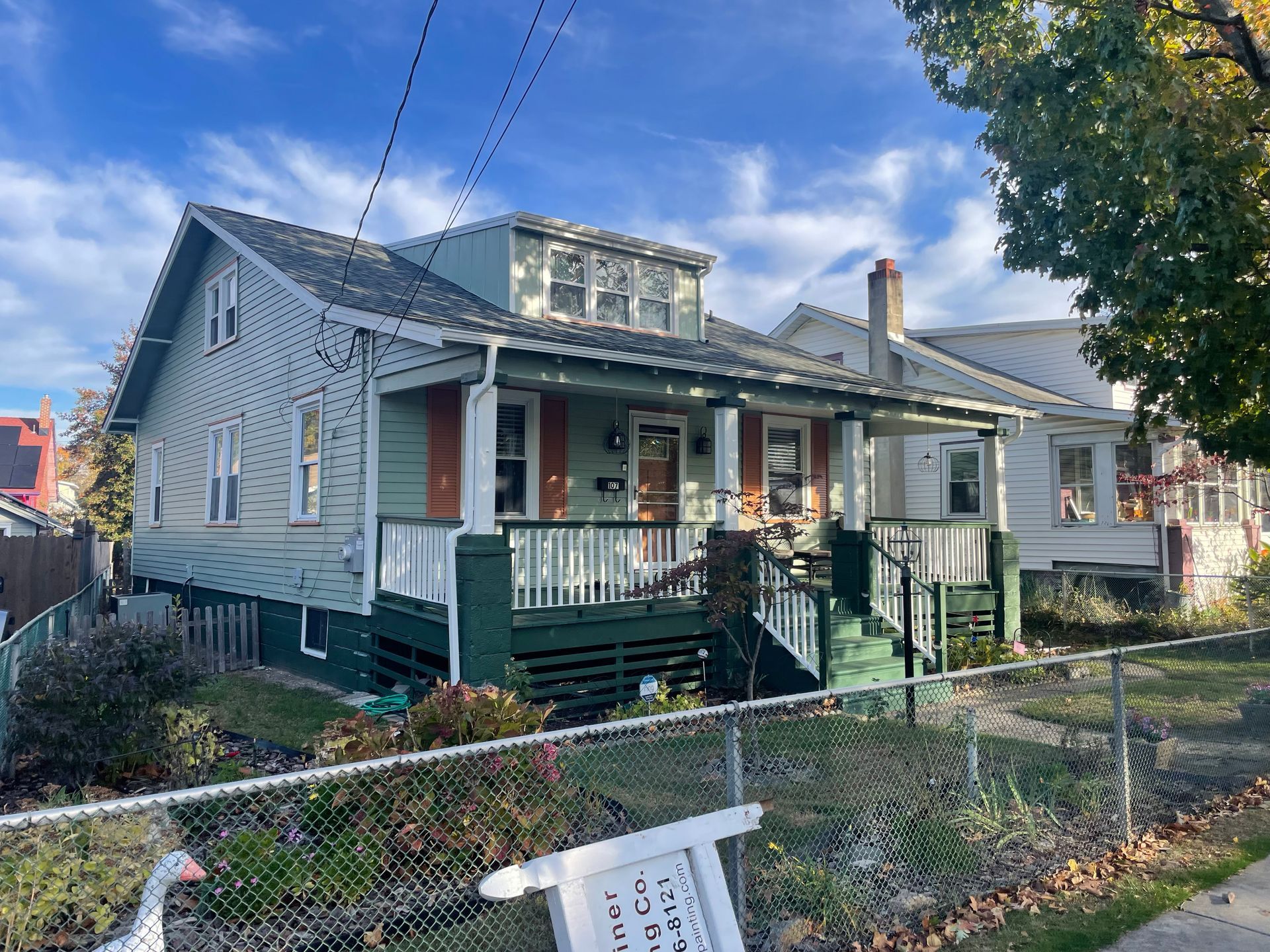 Two-story house with green and white exterior, porch, and a fence in front. Blue sky.