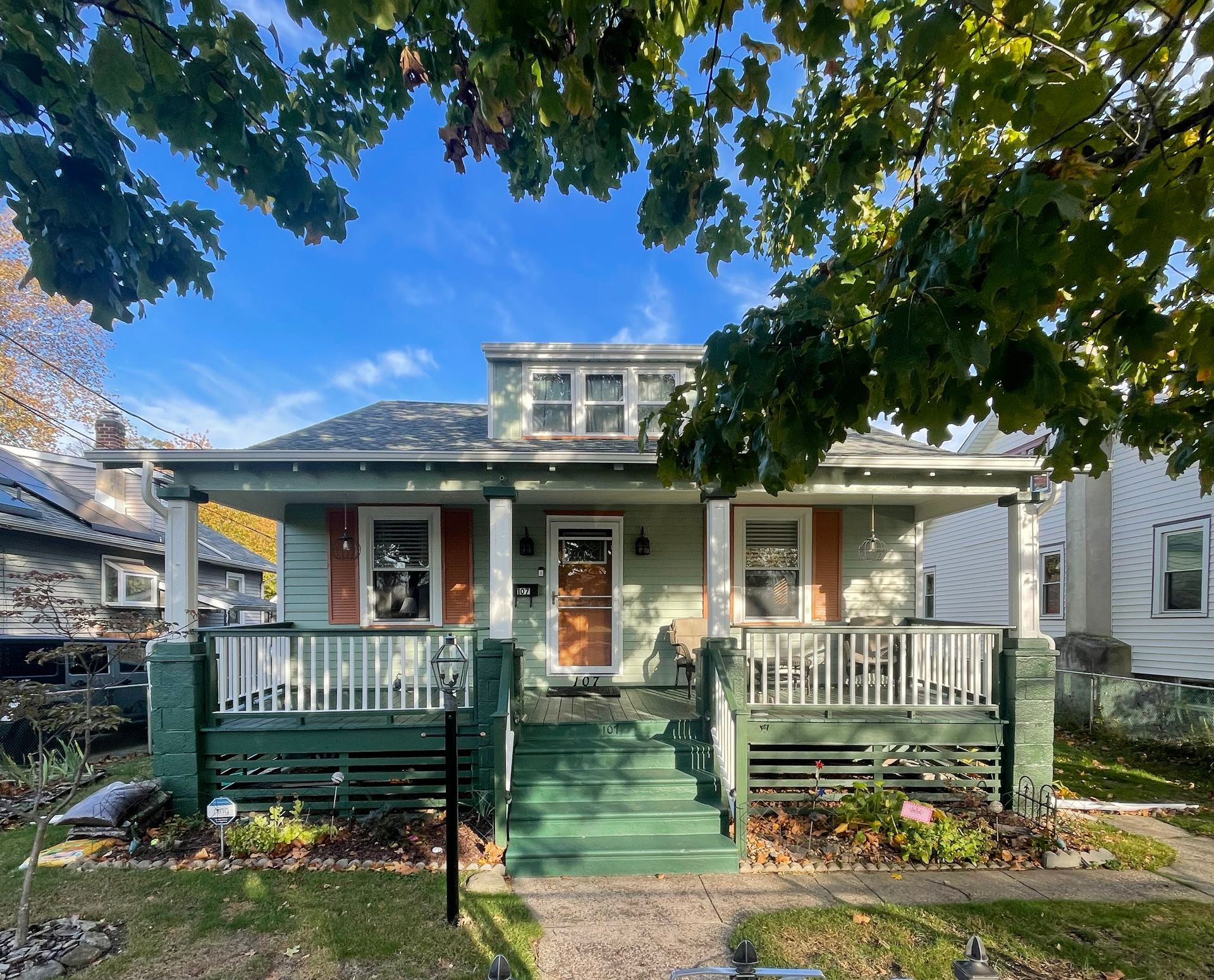 Green bungalow with porch, orange shutters, under a leafy tree, with blue sky.