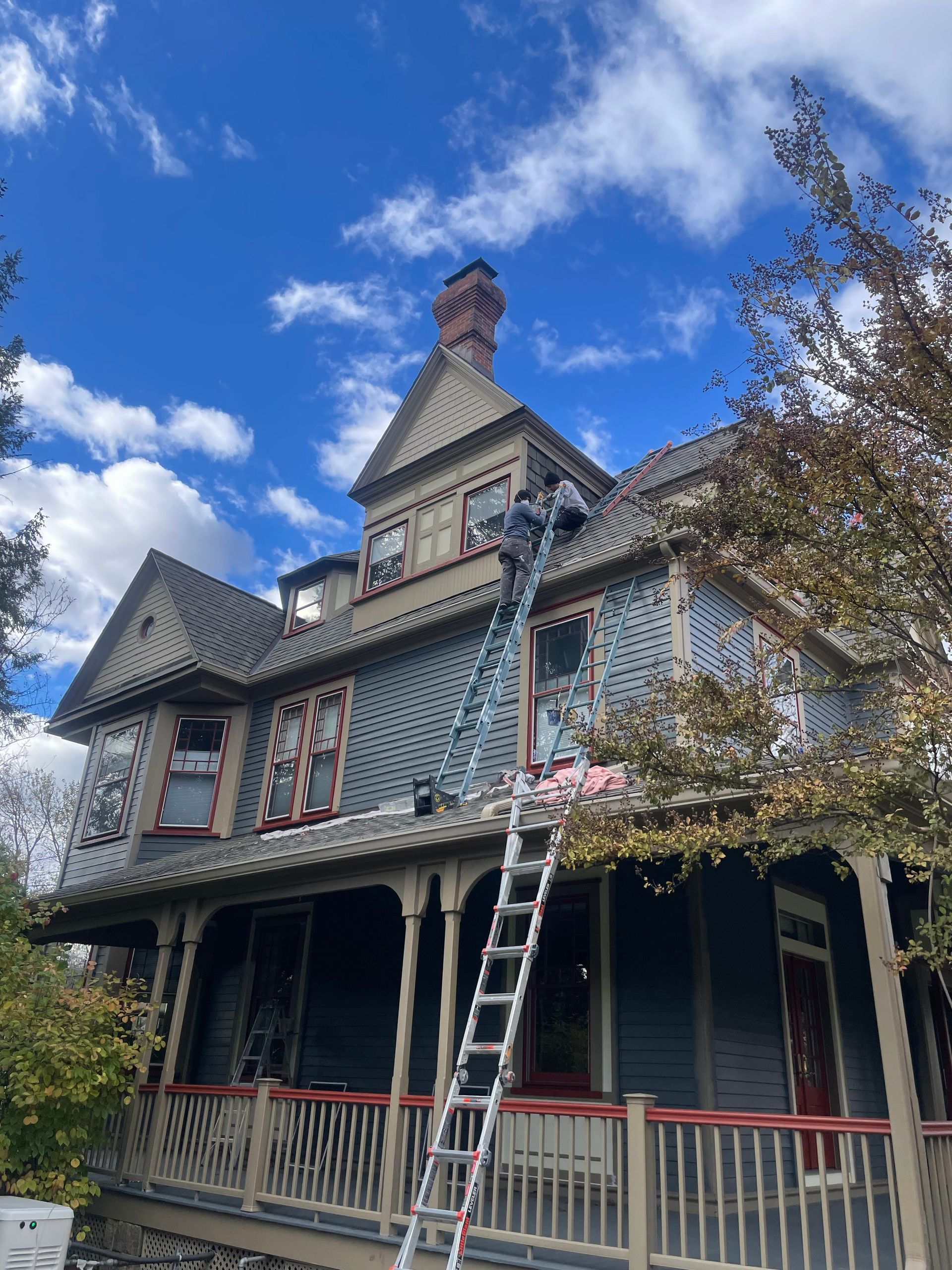 Victorian house with ladder on roof, worker on top. Blue siding, red trim, cloudy sky.