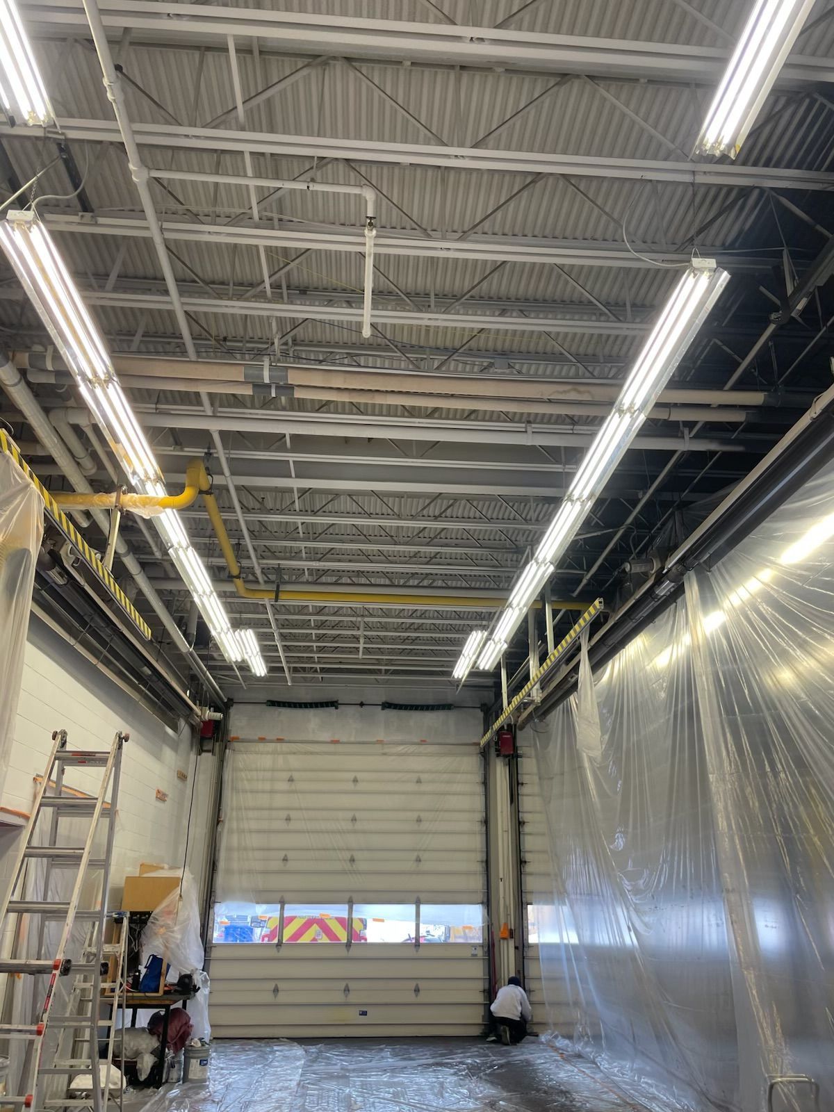Interior view of a garage with an open bay door. Fluorescent lights illuminate the ceiling. A person is visible near the door.