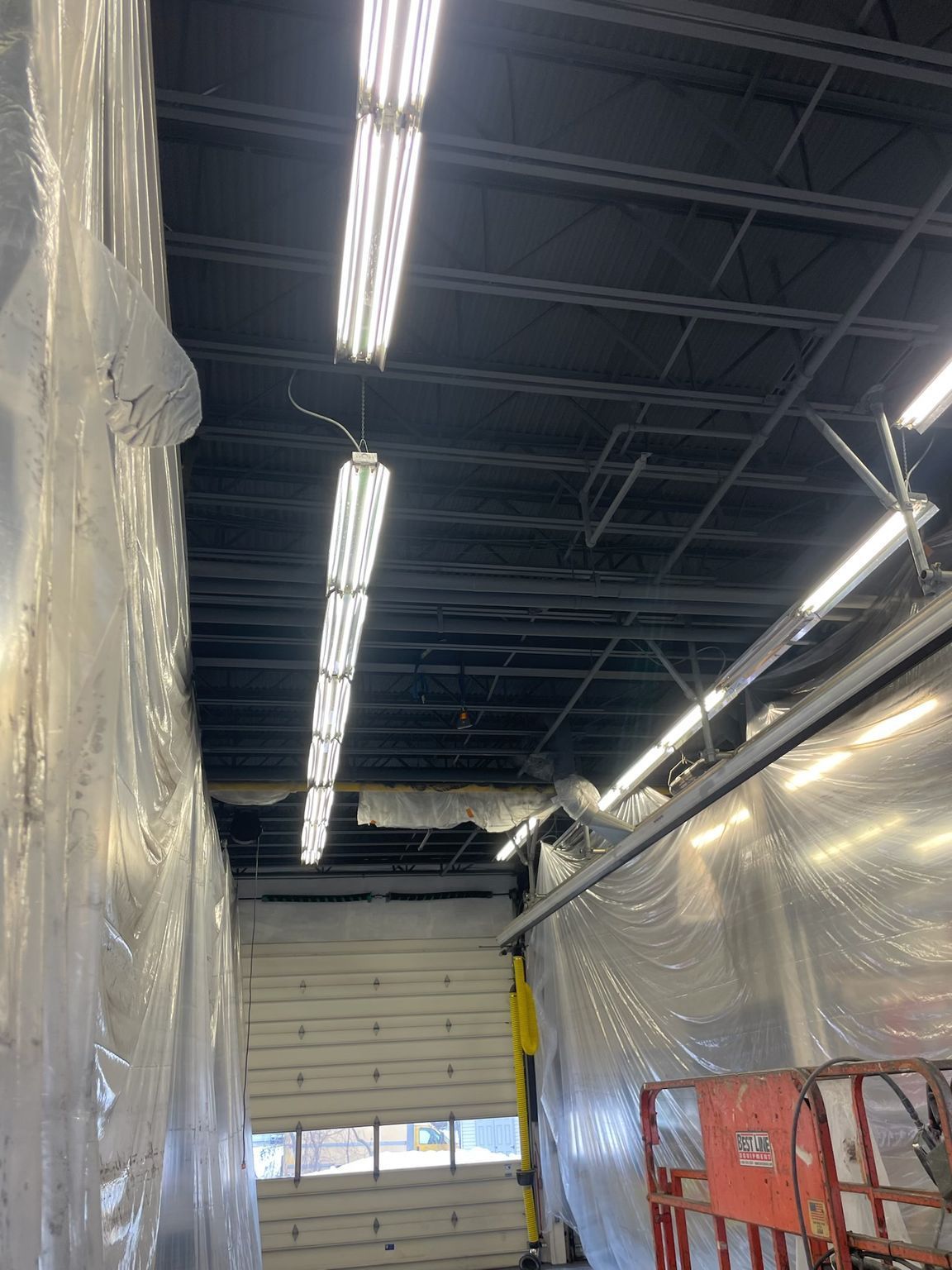 Interior of a garage; fluorescent lights on dark ceiling. Clear plastic covers walls, roll-up door visible.