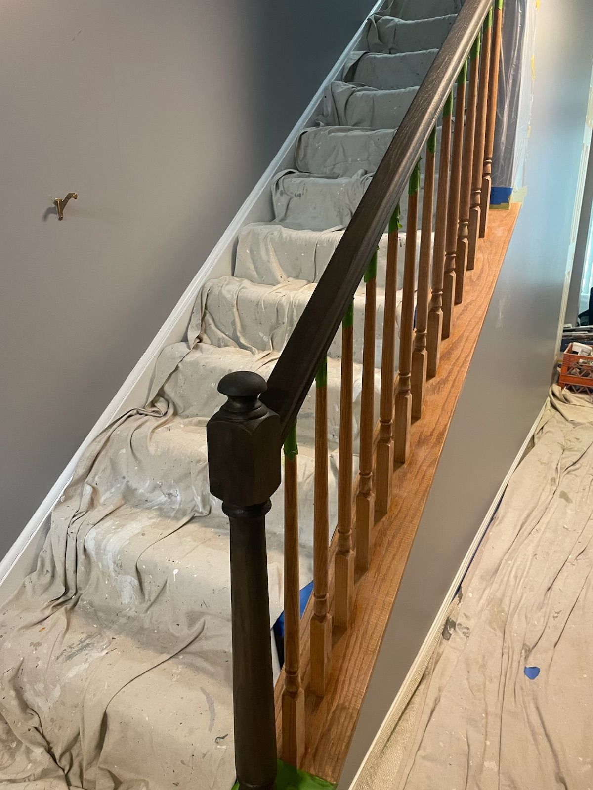 Staircase with brown banister and light wood spindles. Steps covered in drop cloths, grey walls.