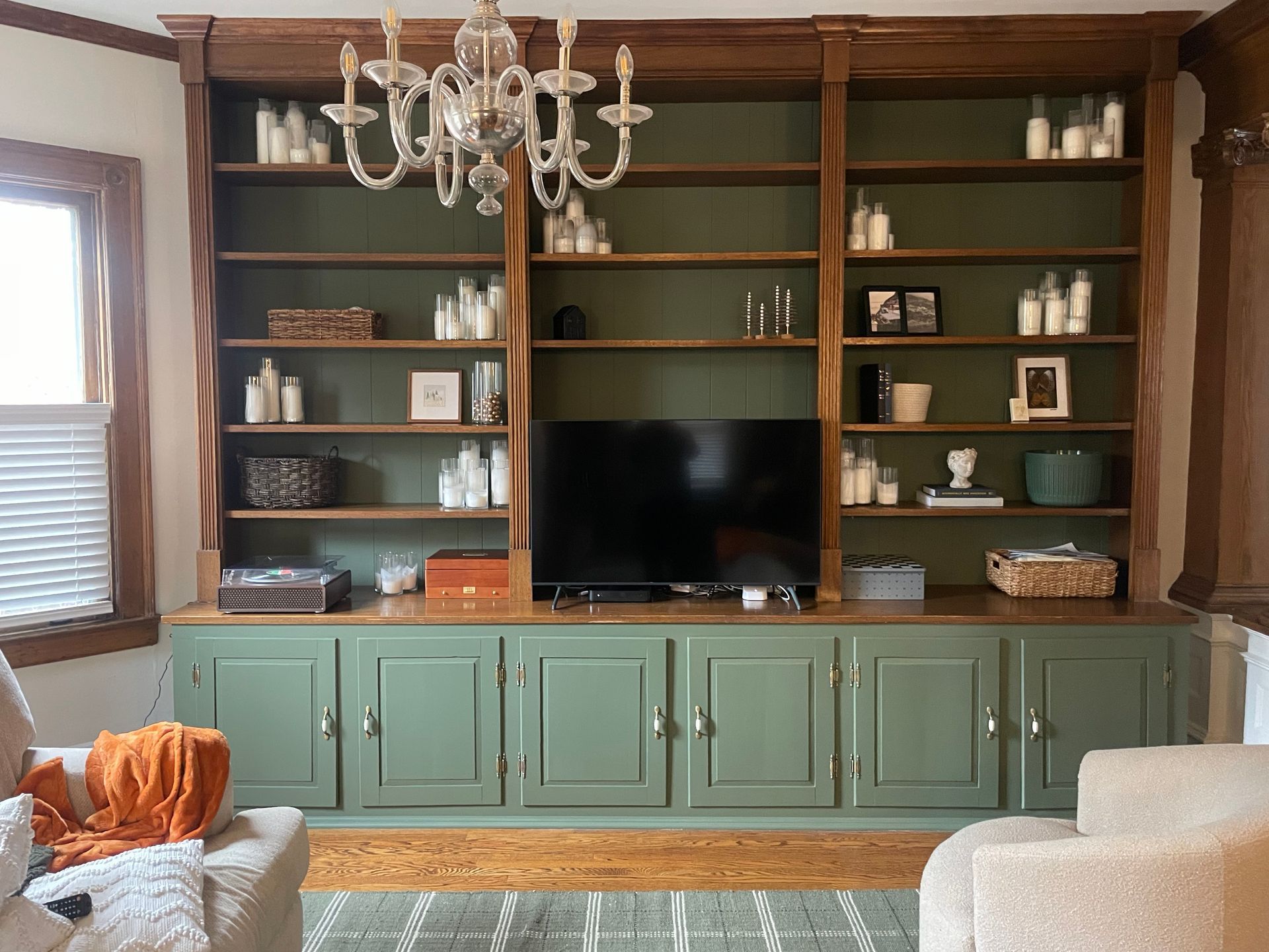 Built-in bookcase with green cabinets, shelves lined with candles and decor, TV in the center, and a chandelier.