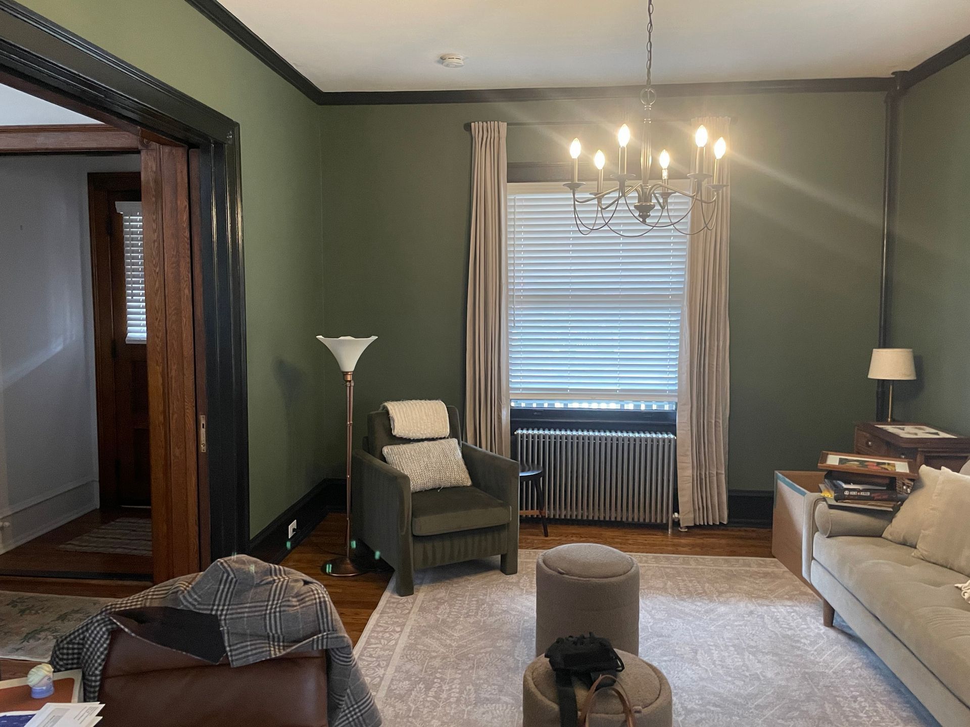 Green-walled living room with a reading chair, sofa, window with blinds, and chandelier.