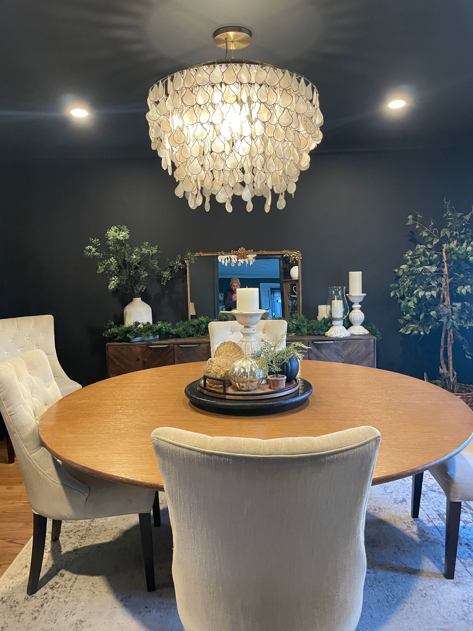 Round wooden dining table with chairs, under a beaded chandelier, against a dark gray wall.