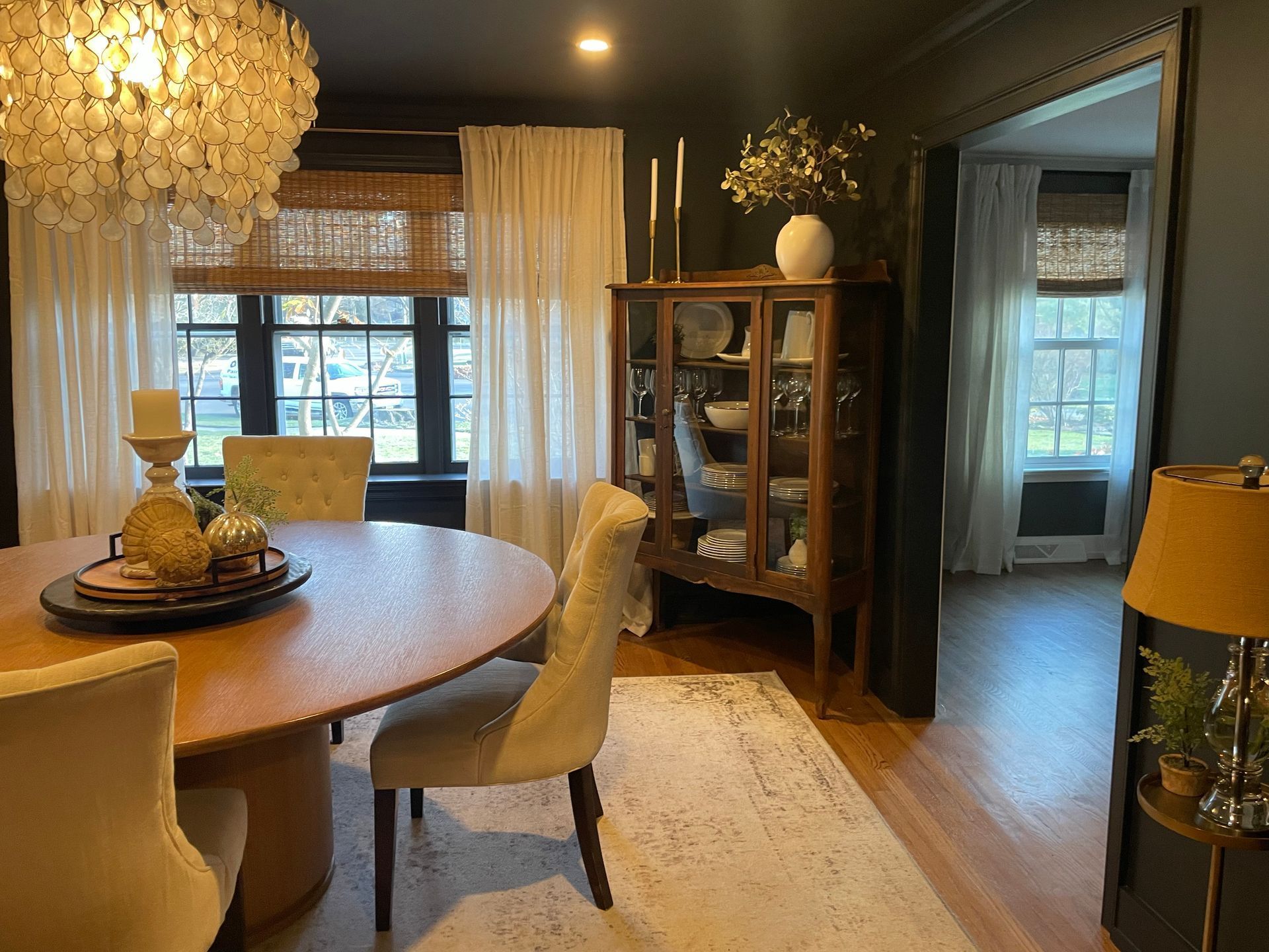 Dining room with wood table, upholstered chairs, glass cabinet, and curtains; dark walls, gold chandelier.