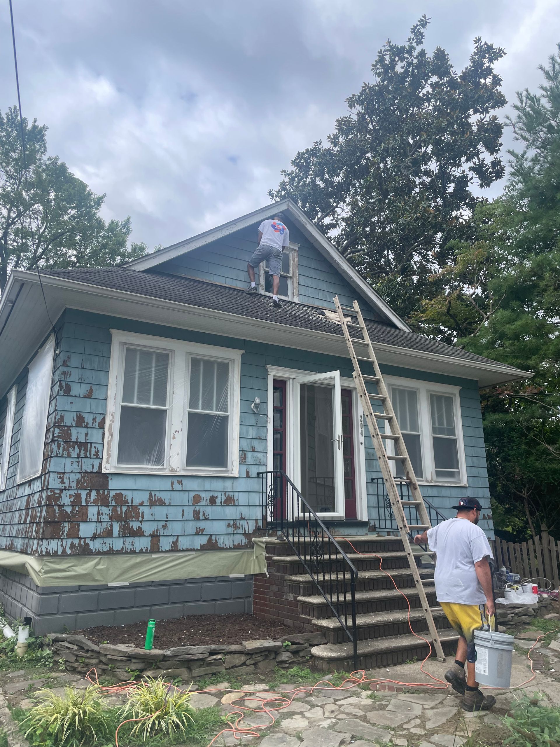 Workers painting a light blue house. One on a ladder, another on the roof. Cloudy sky.