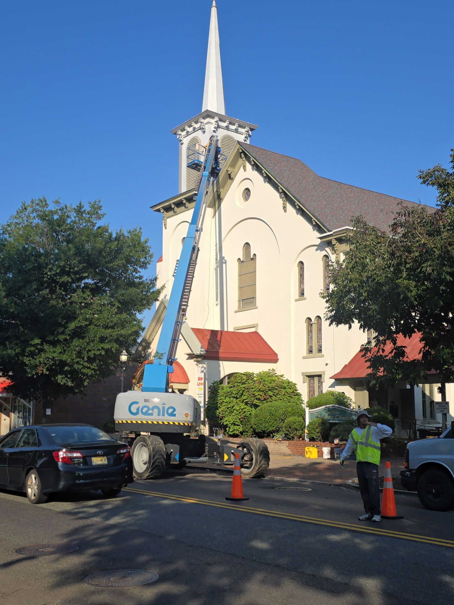 A Genie lift reaching toward a church spire; worker directs traffic, cars parked on street.