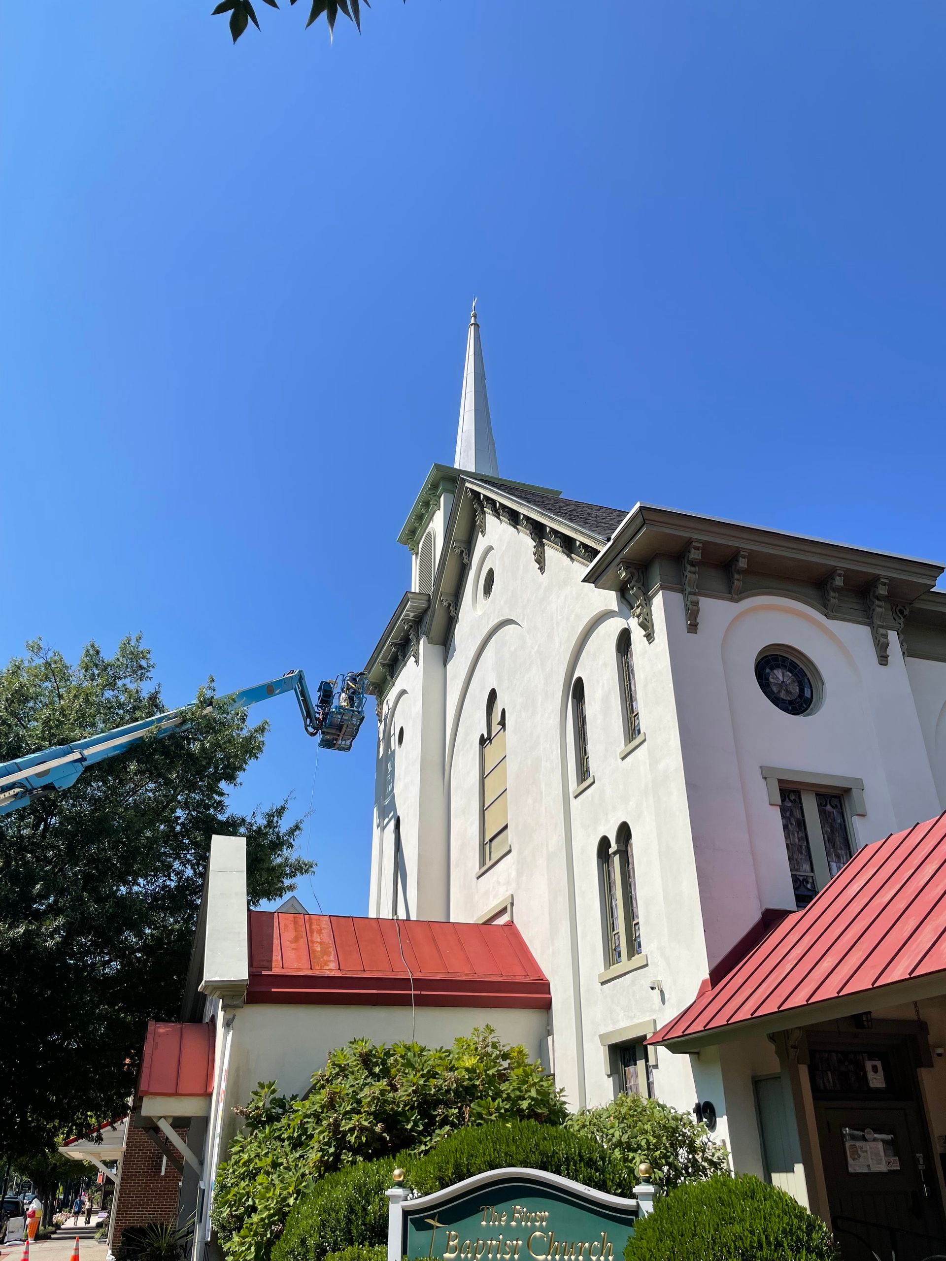 White church with tall spire under blue sky. A lift is used for construction. Red roof and green bushes.