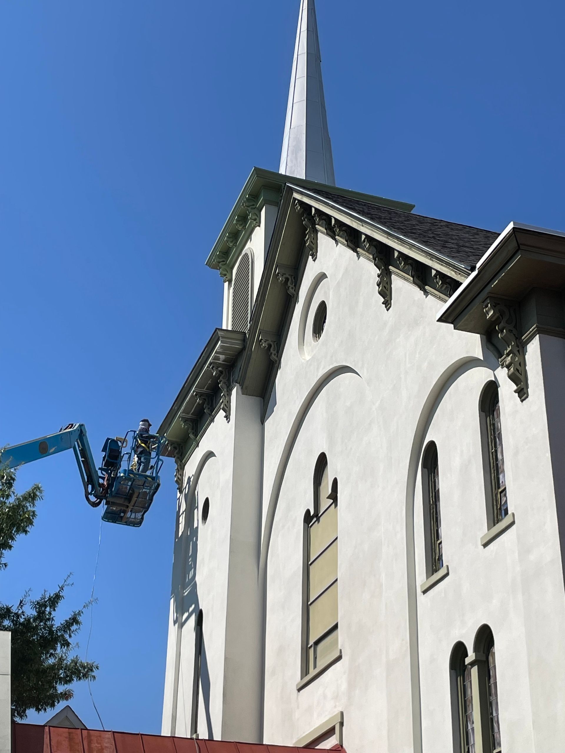 A person in a blue lift working on the exterior of a white church with a tall steeple against a blue sky.