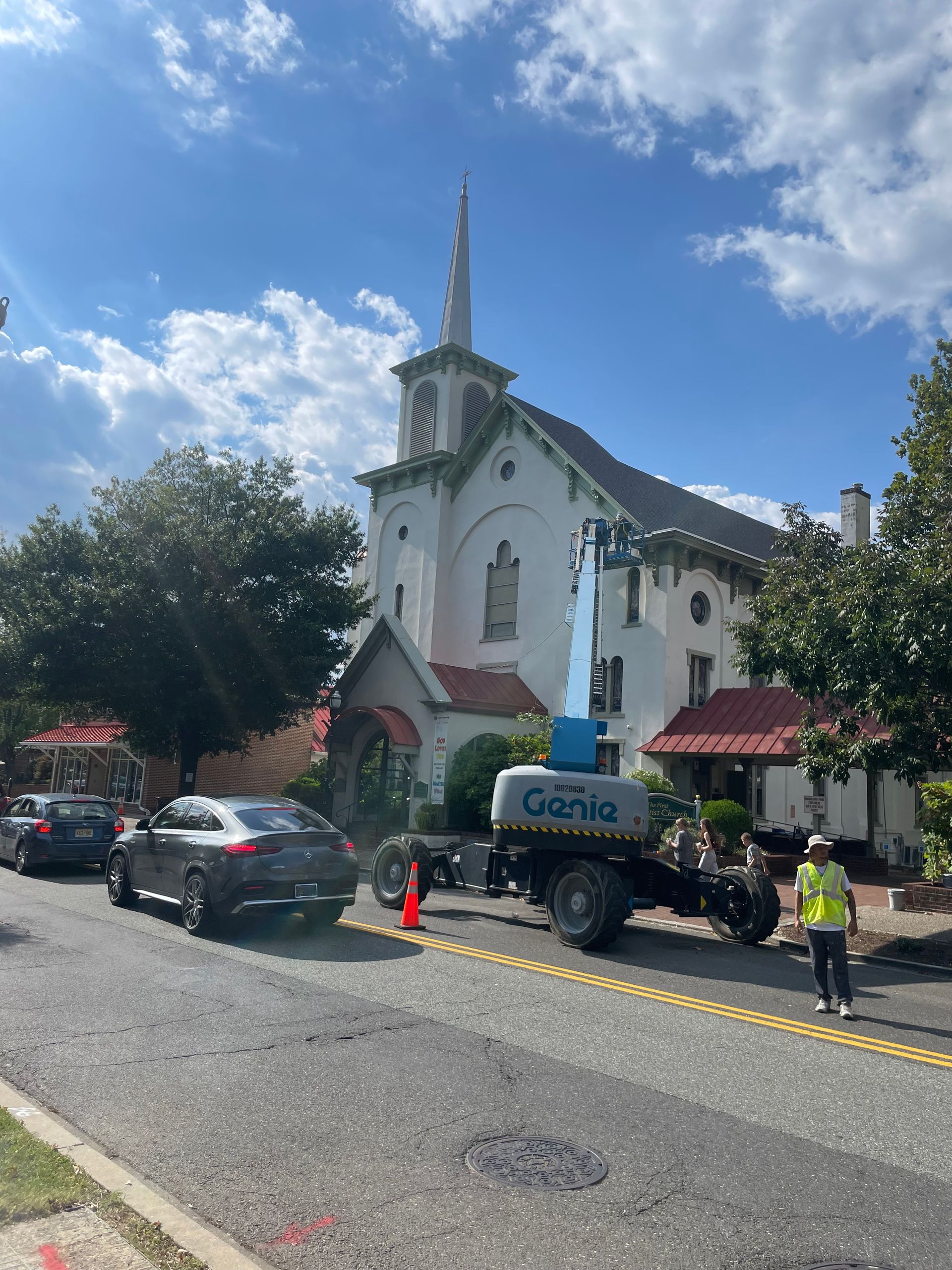 A blue Genie lift in front of a white church with a steeple; a worker in a safety vest stands nearby.