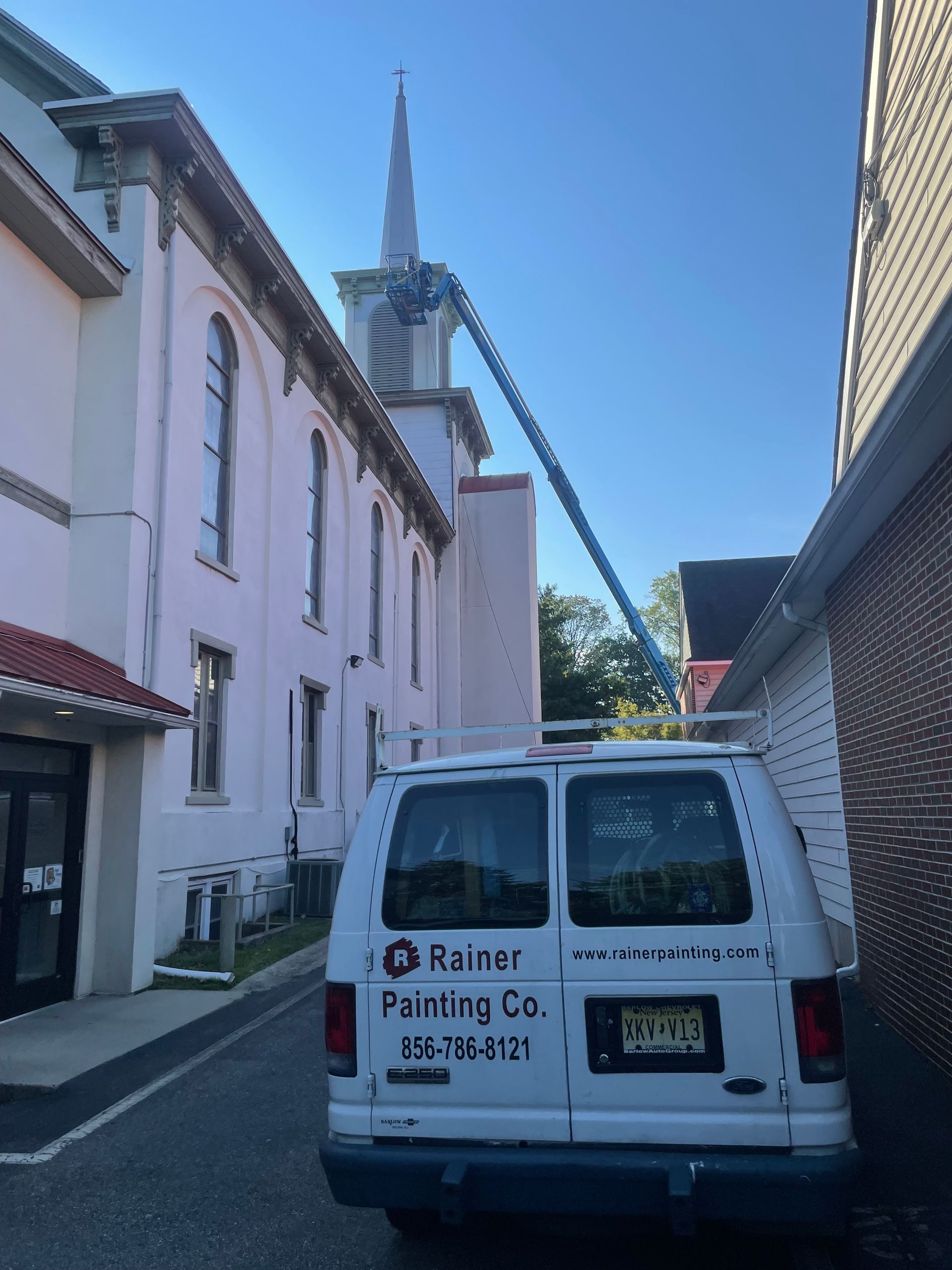 White van parked by a church. A blue lift extends to the church steeple for painting.
