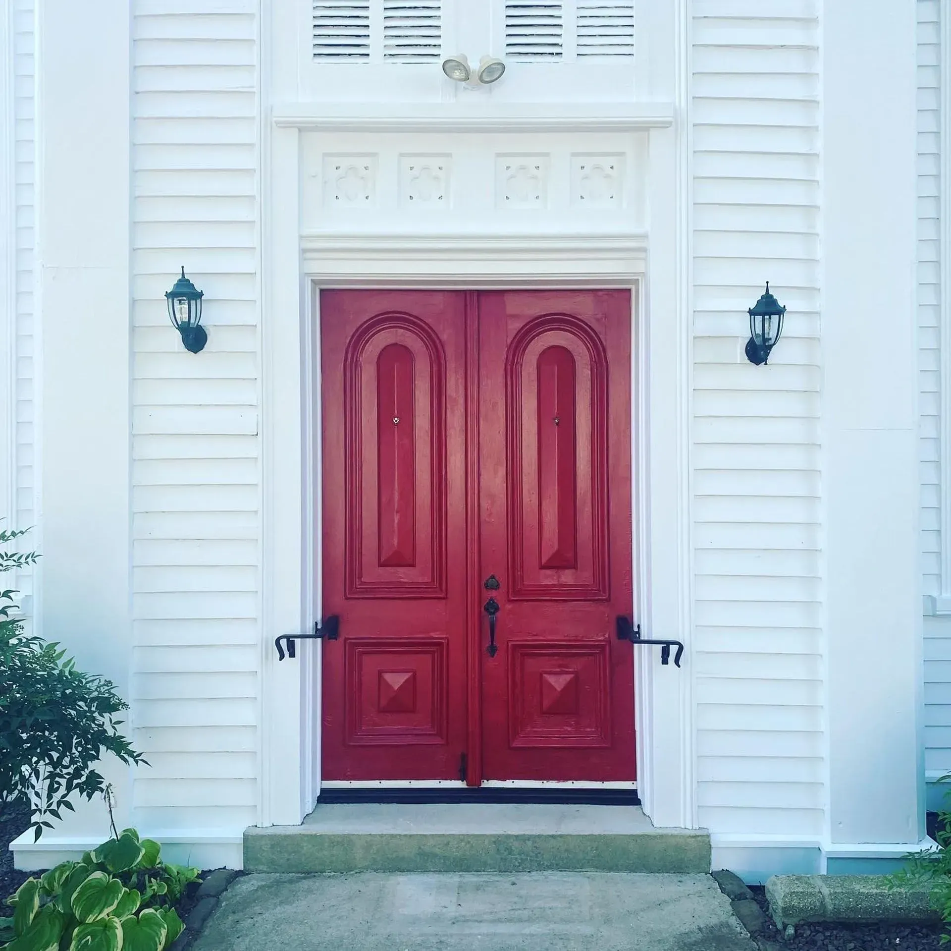 Red double doors centered on a white building with sidelights, sconces, and porch.