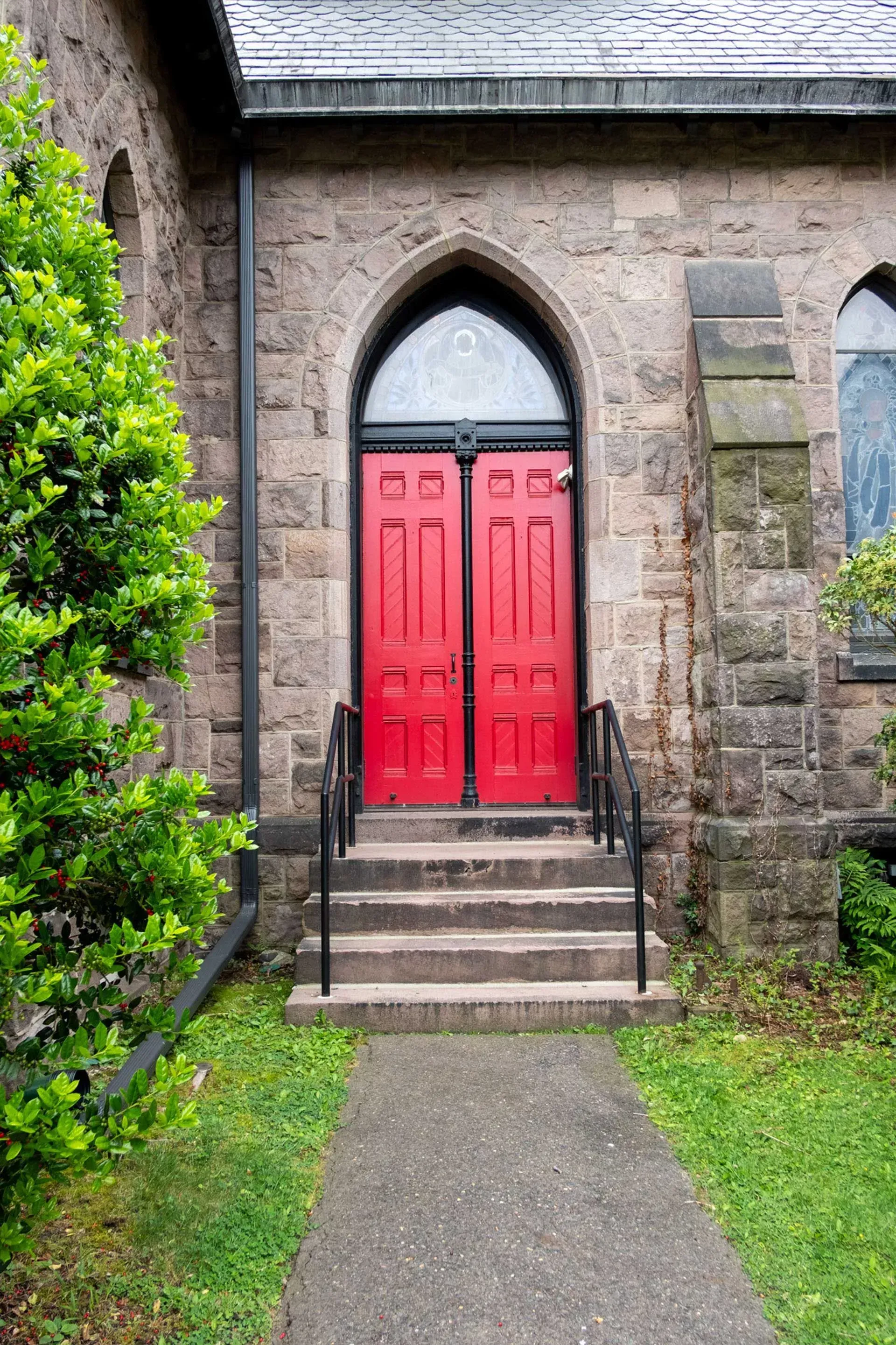 Red double doors of a stone church with arched top, small steps, and a pathway.