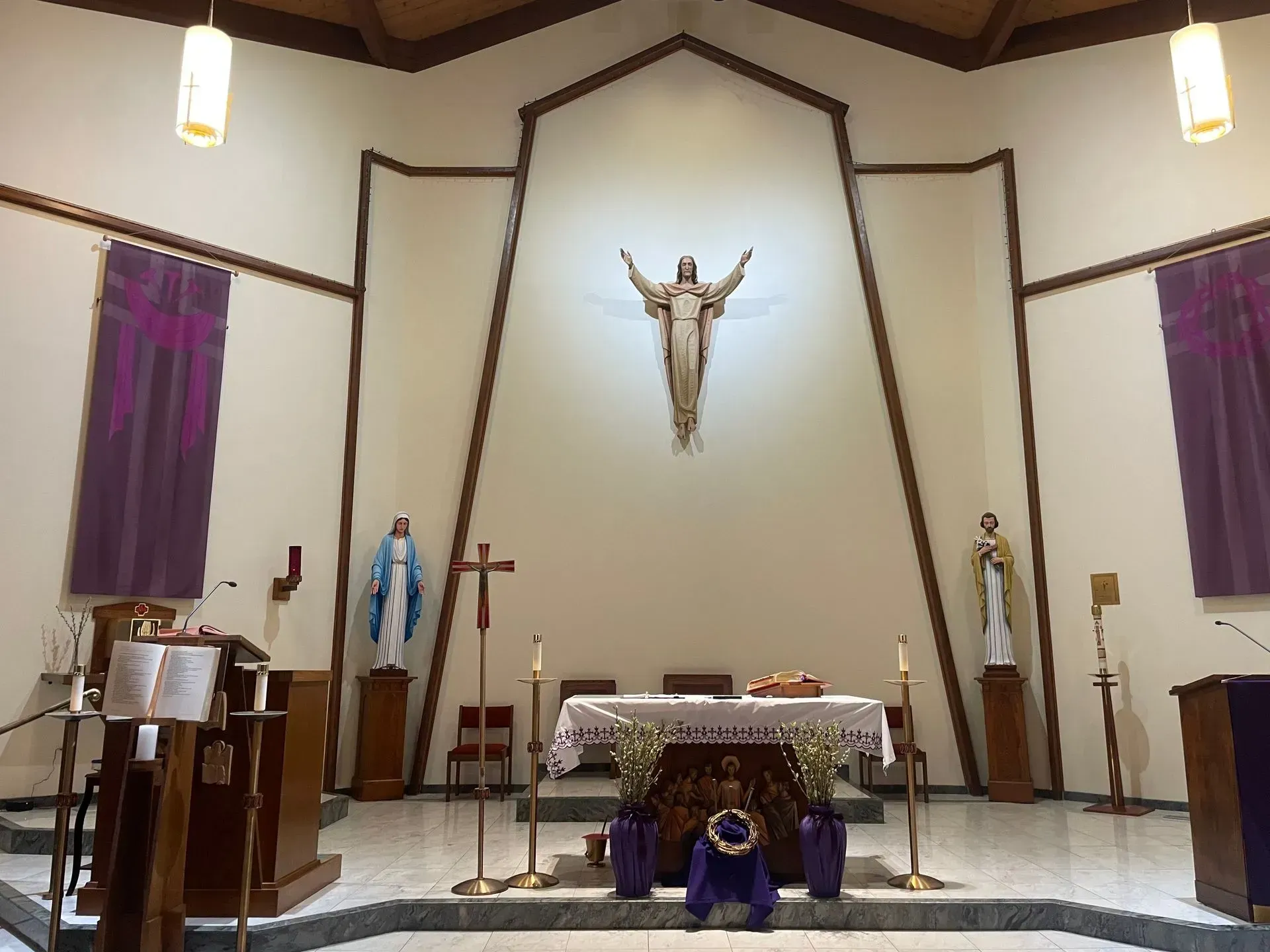 Church interior with altar, statue of Jesus, statues of Mary and a saint. Purple curtains.