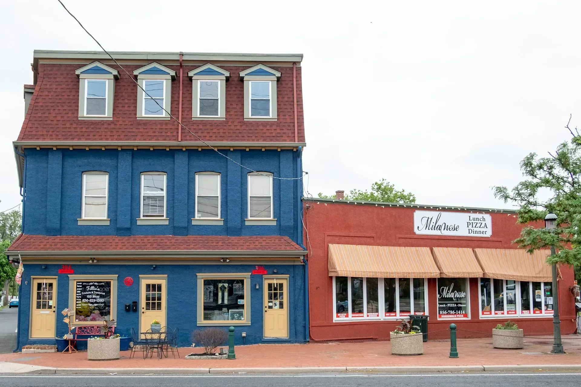 Blue and red buildings with storefronts on a brick sidewalk. Yellow doors, awnings, and dormers visible.