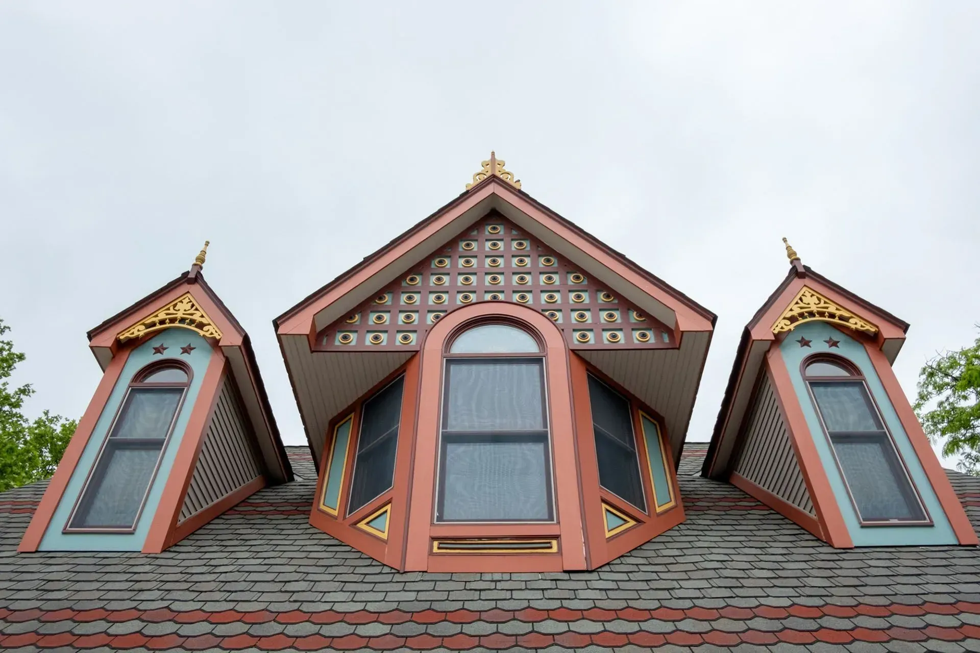 Three ornate, dormer windows with arched windows and gold accents on a gray roof under a cloudy sky.
