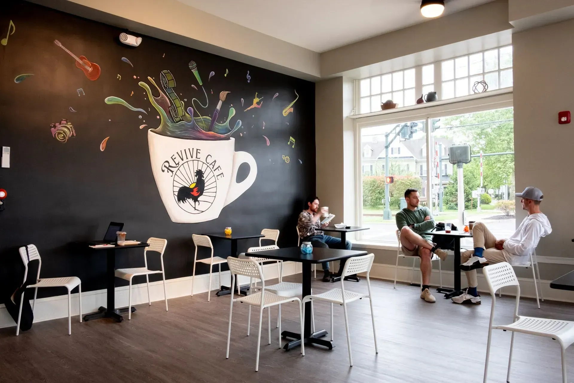 Coffee shop interior with mural on black wall, tables, chairs, and people near window.