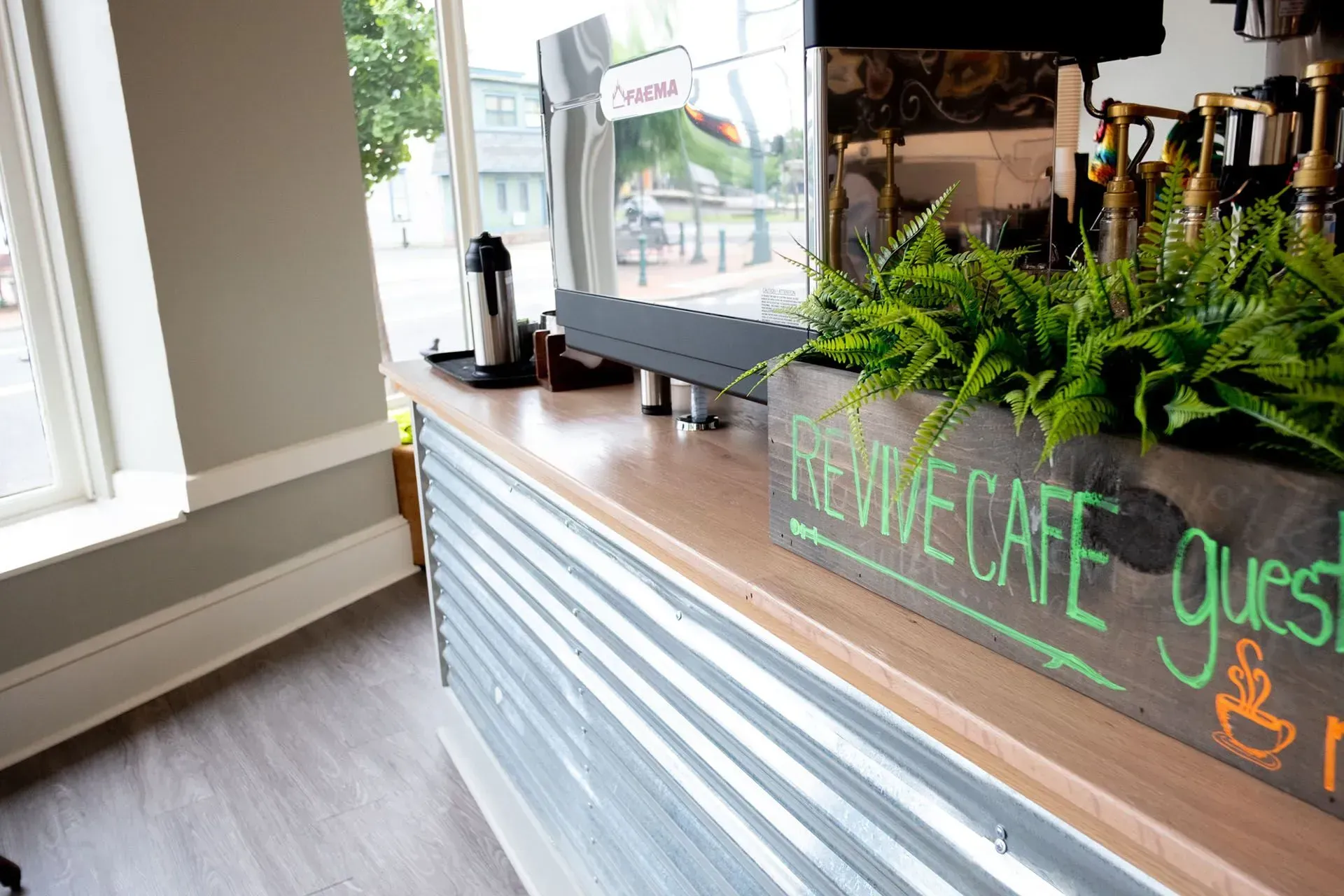 Coffee shop counter with metal paneling, wooden top, and plants; a sign reads 