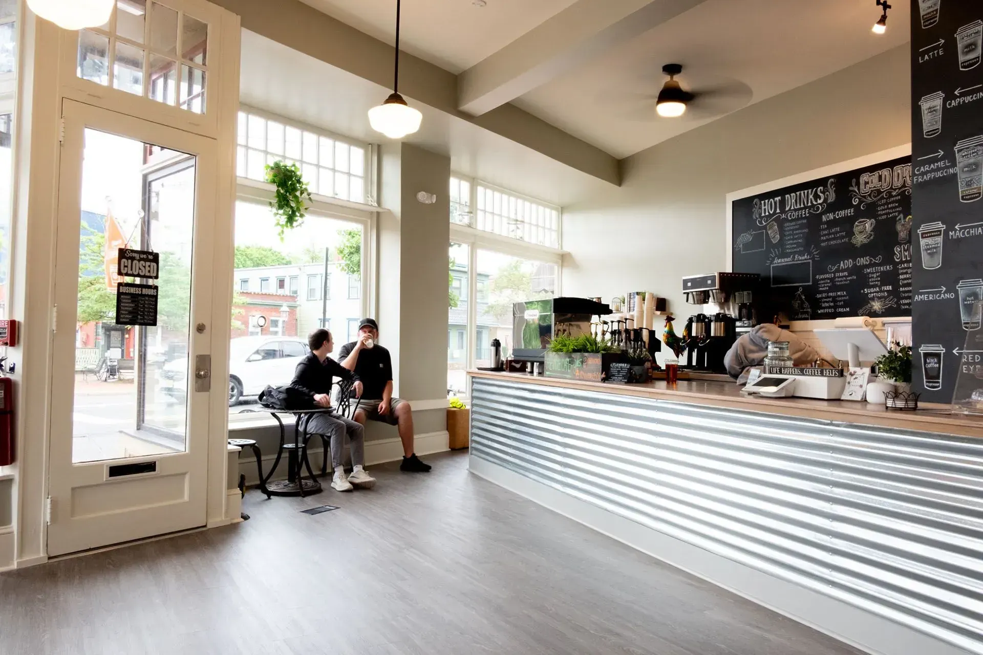 Coffee shop interior with two people sitting near window, counter, and menu board.