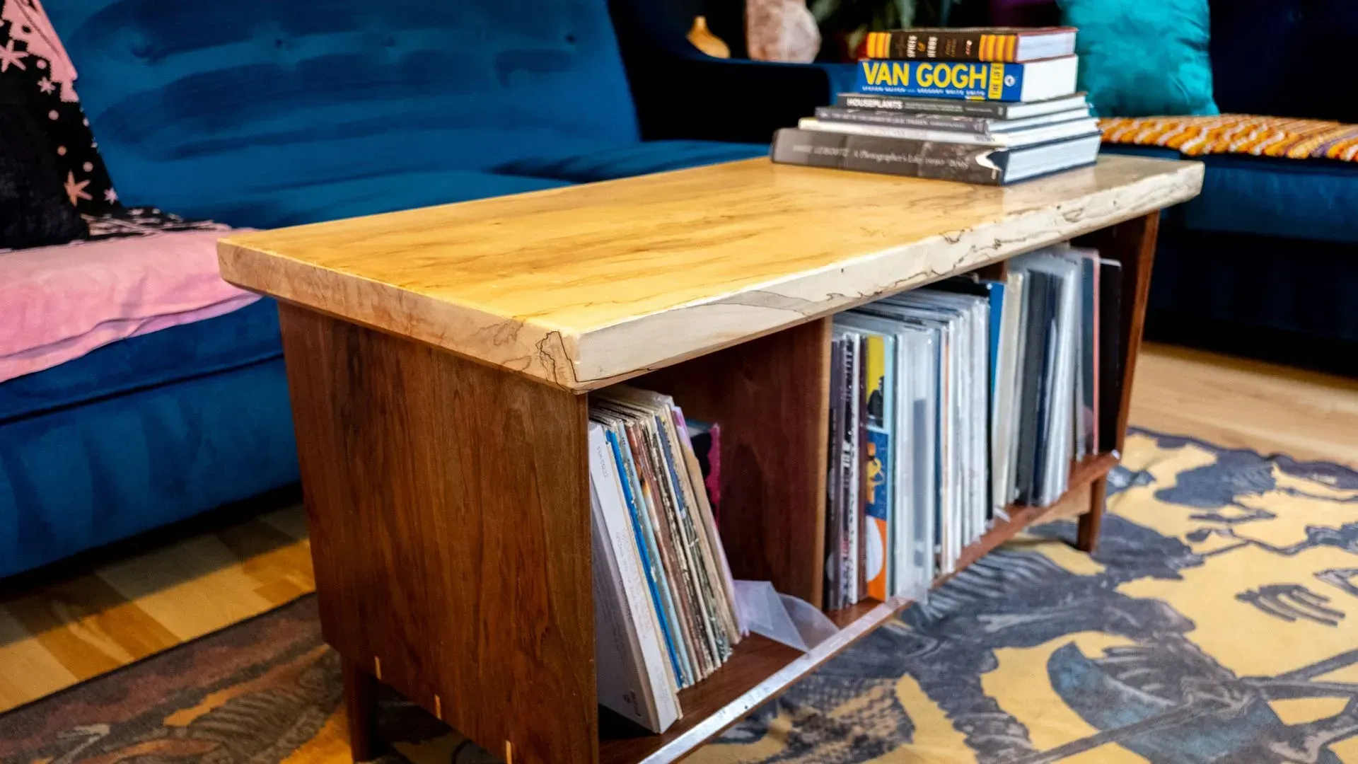 Wooden coffee table with books on top and in open storage. Teal couch in background.