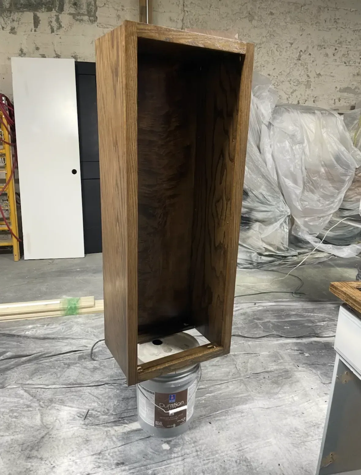 Wooden box stained brown, sitting on a paint bucket in a workshop.