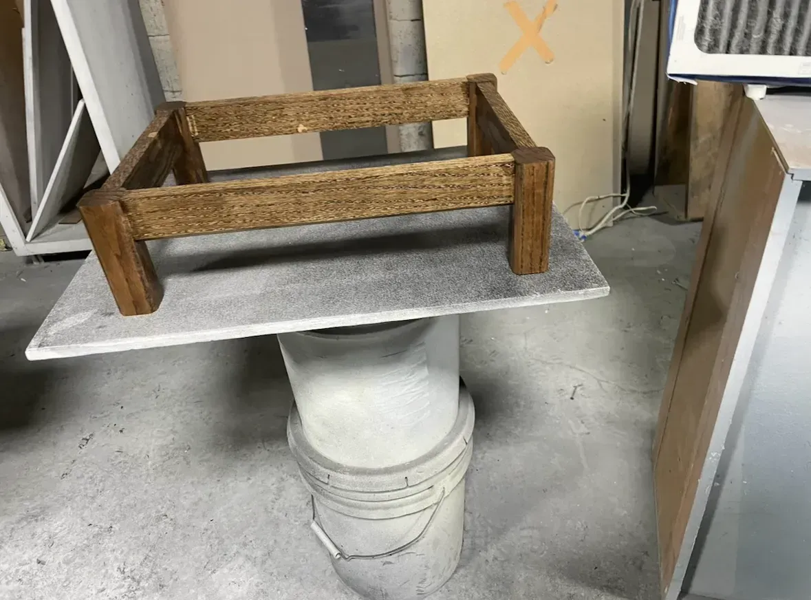 Wooden frame on a gray square tile, resting on a white bucket, in a workshop.