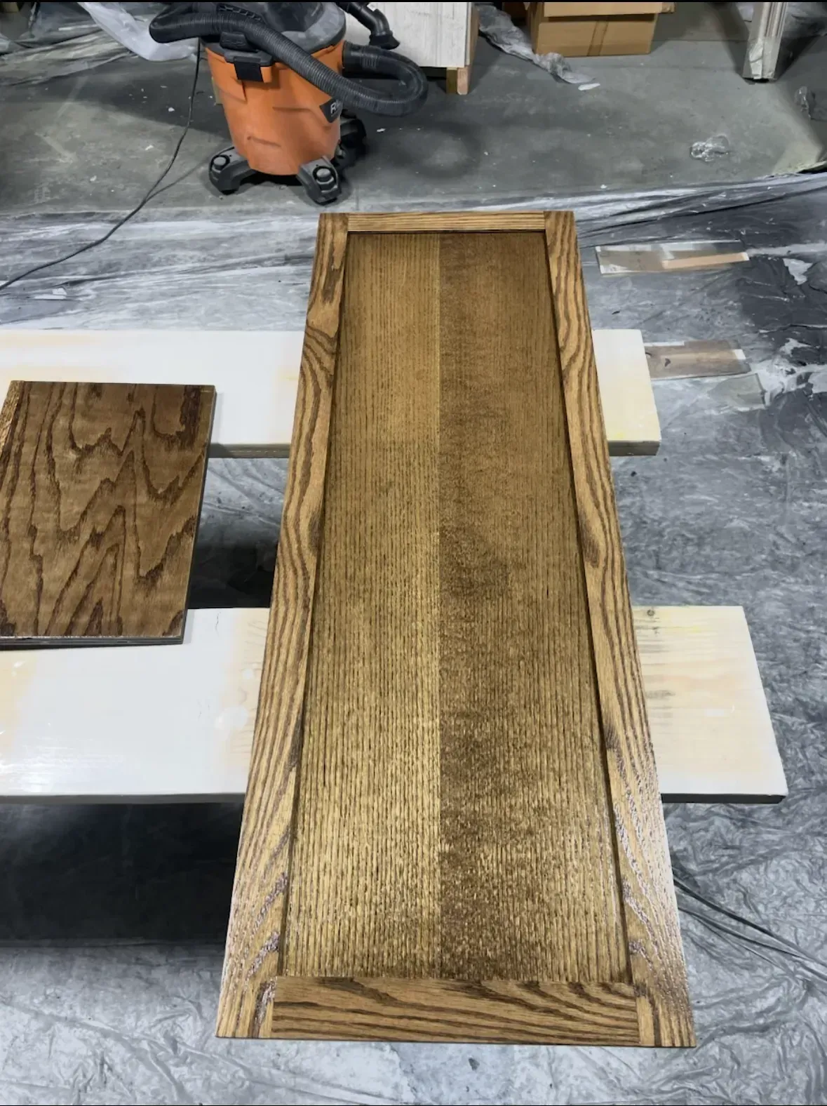 Wood cabinet door and small panel stained brown, on white boards, shop setting with vacuum.