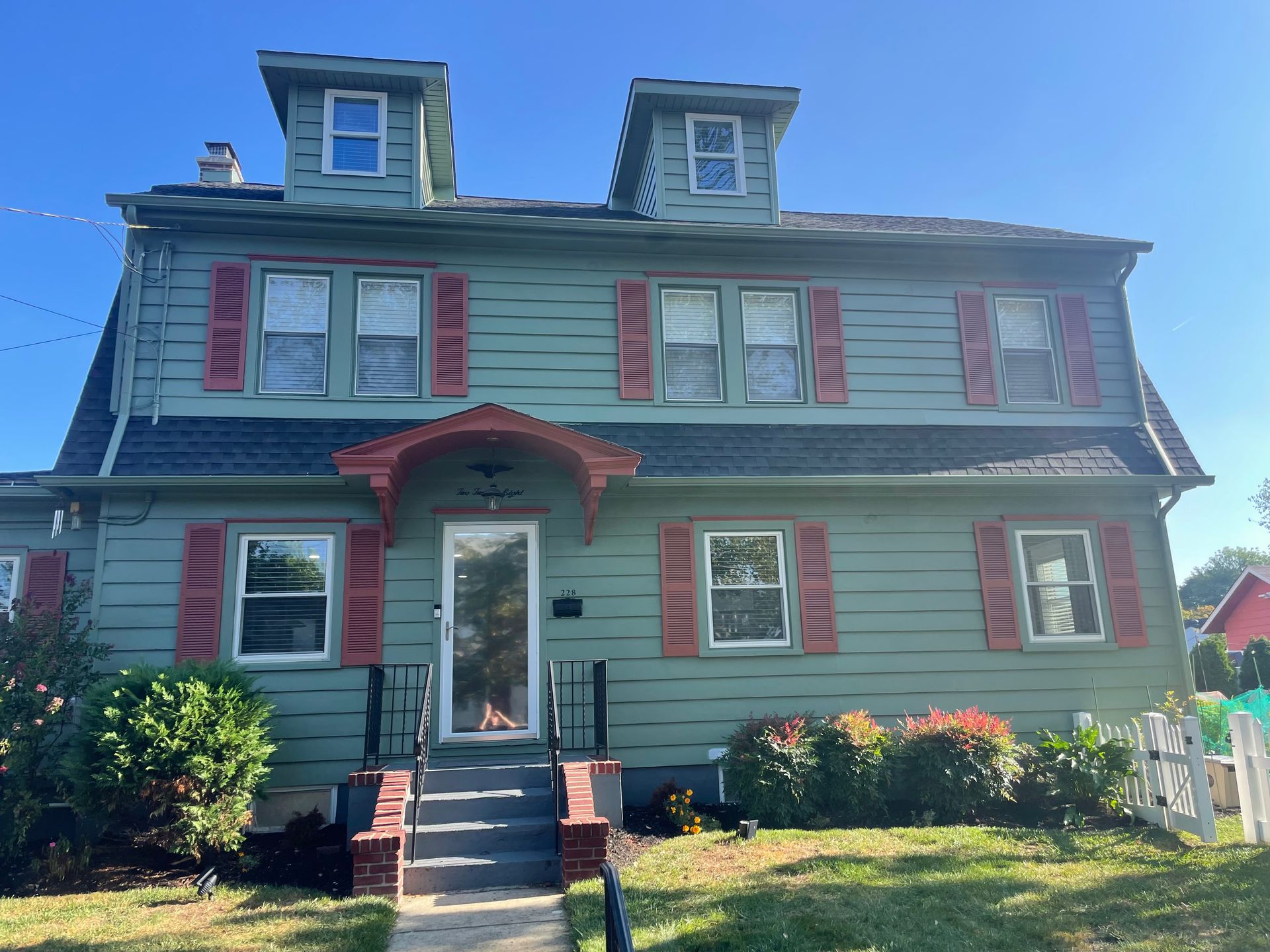 Green house with red shutters, two dormers, and a small front porch on a sunny day.