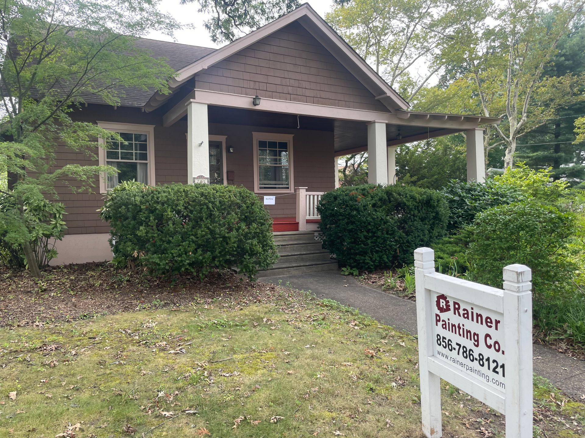 Brown house with a porch, fronted by bushes, with a Rainer Painting Co. sign in the yard.