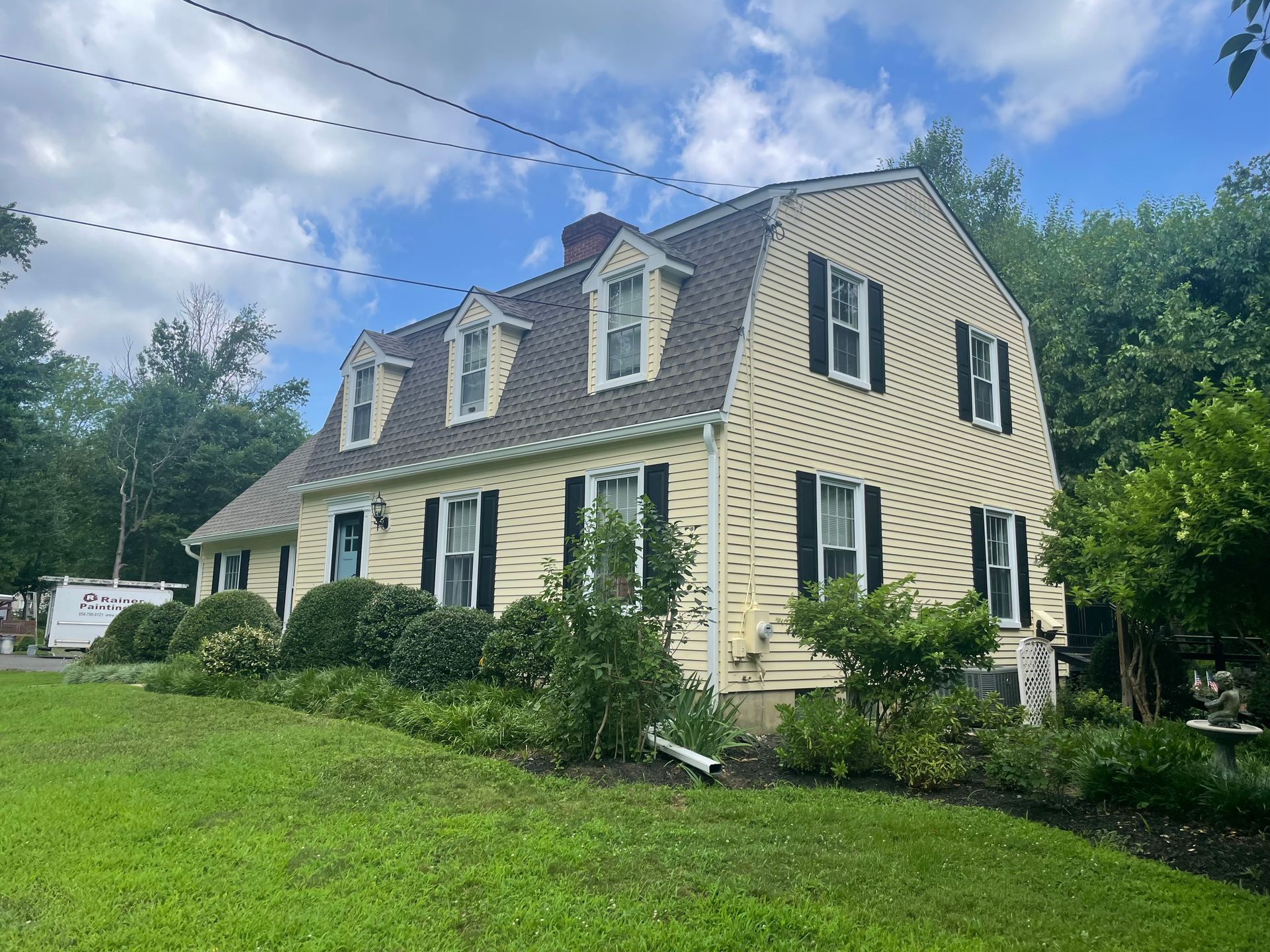 Yellow house with black shutters, three dormers, and a green lawn. Cloudy sky in the background.