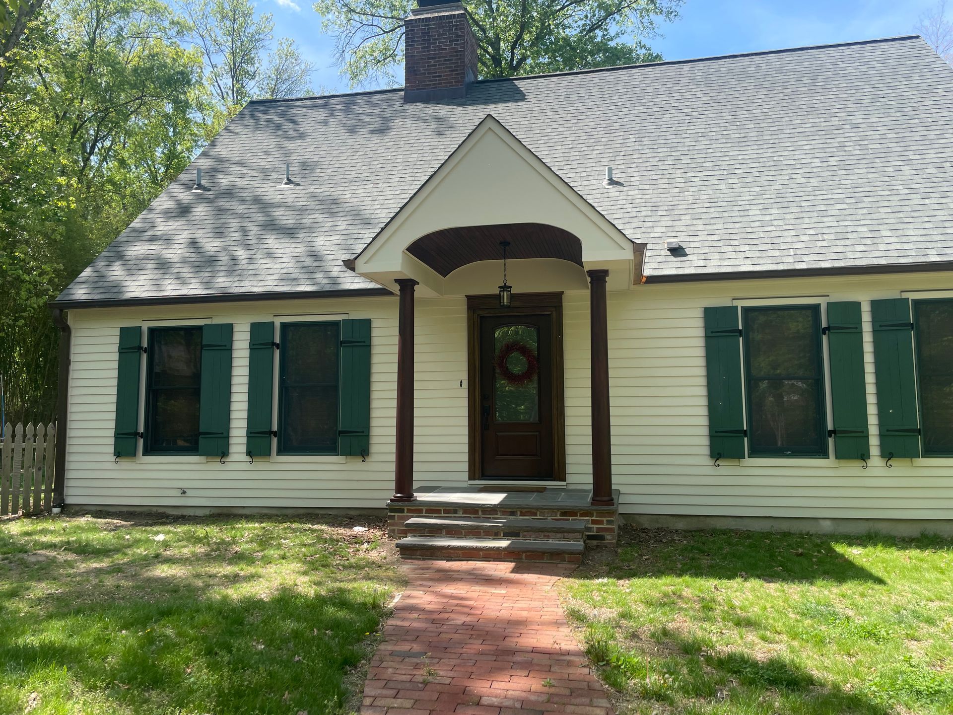 White house with green shutters, brown door, brick walkway, and a small porch.