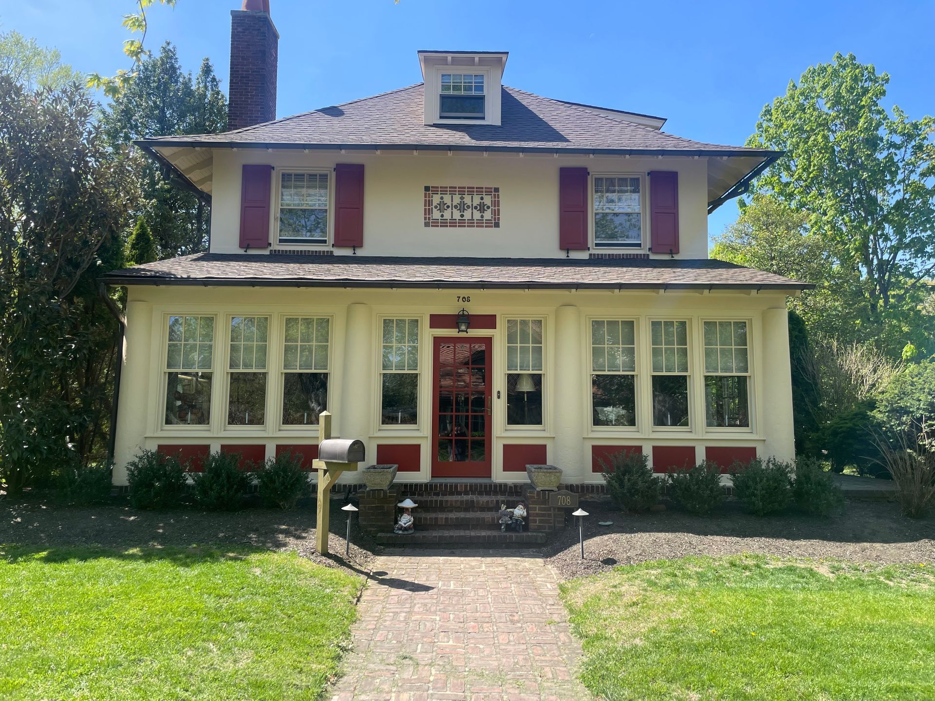 Two-story house with beige siding, red shutters and trim, brick walkway, and a green lawn.