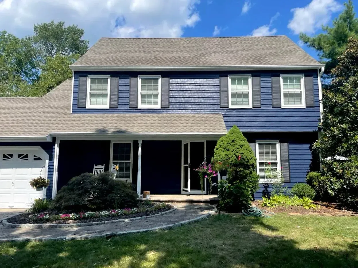 Two-story blue house with white trim, gray roof, and landscaping on a sunny day.