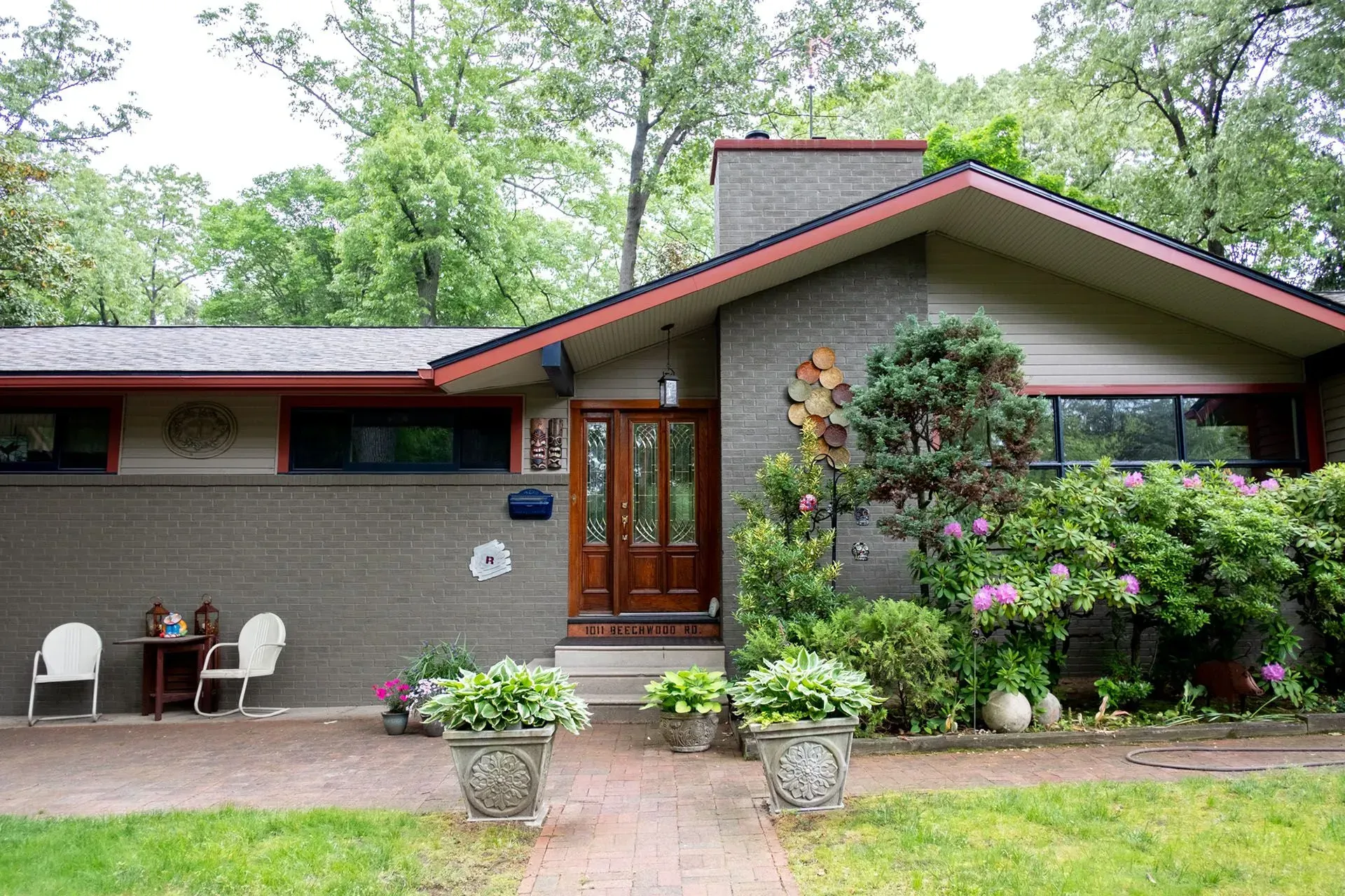 Ranch-style house with brick facade, red roof, double wood doors, surrounded by greenery and potted plants.