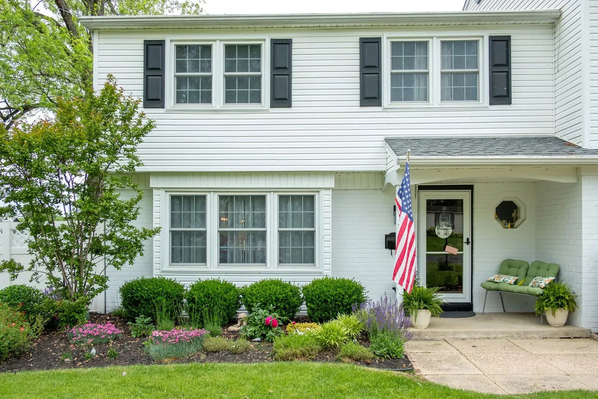 White two-story house with black shutters, American flag, and front yard garden with green bushes and colorful flowers.