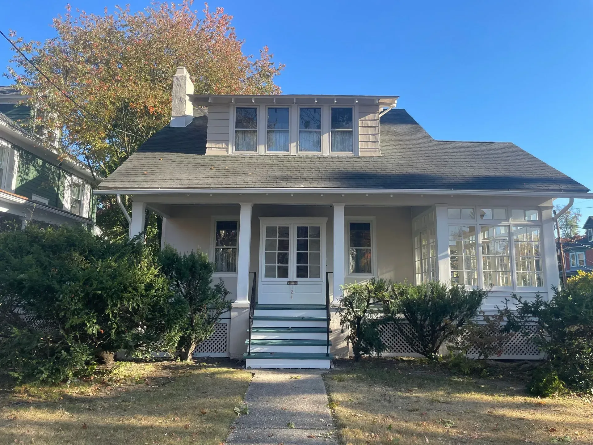 Tan bungalow with porch, steps, and windows. Green bushes in front; fall foliage above.