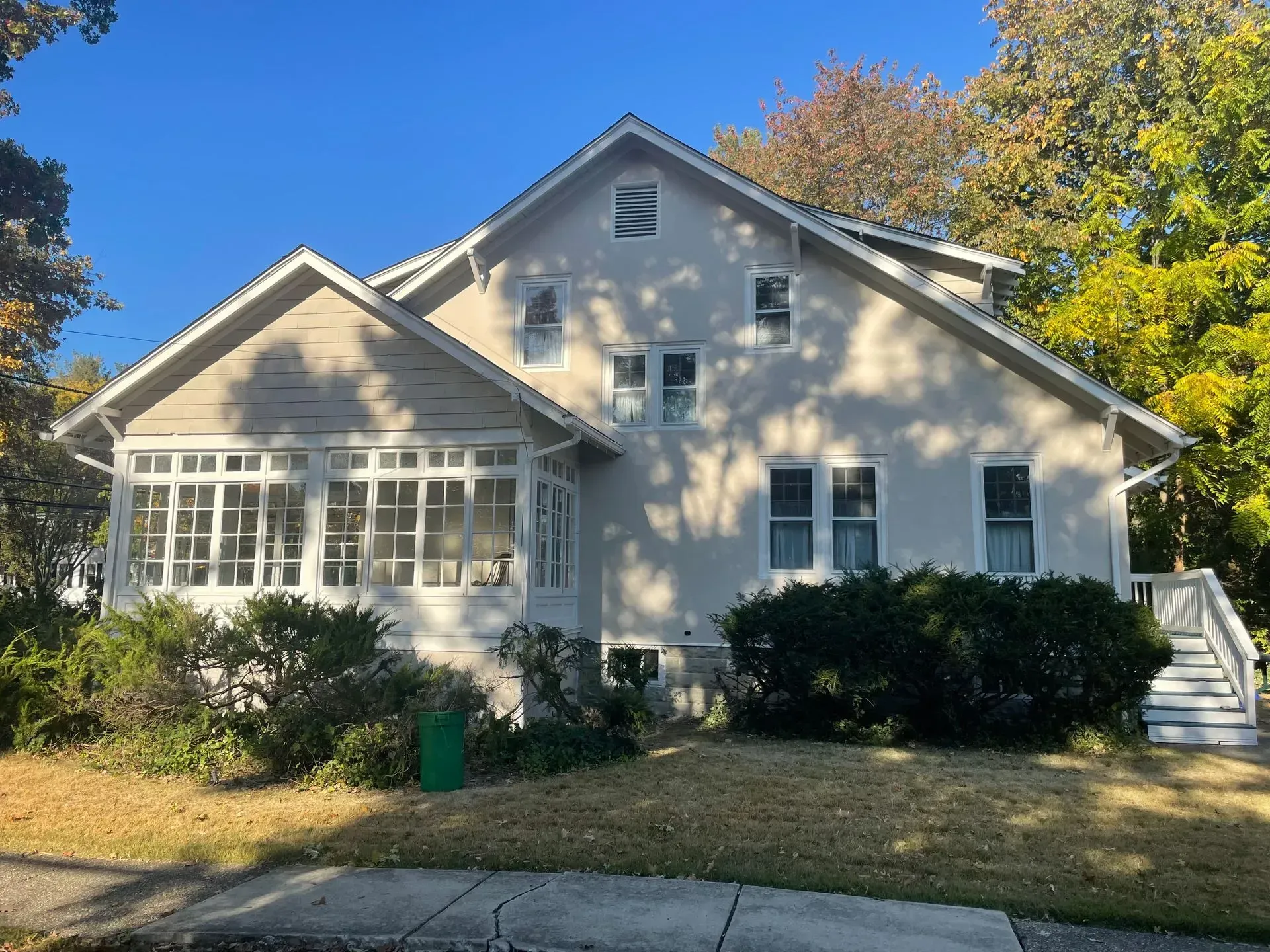 Two-story light stucco house with sunroom, surrounded by trees and shrubs on a sunny day.