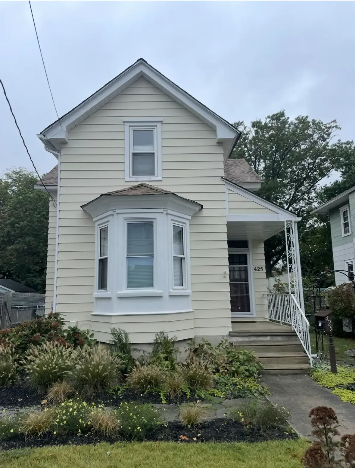 Cream-colored two-story house with a bay window, porch, and landscaping under a cloudy sky.